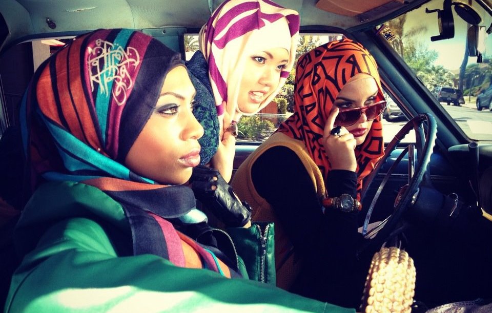 L'image montre trois femmes à l'intérieur d'une voiture. Elles portent des hijabs colorés avec des motifs variés. L'une d'elles est au volant, tandis que les deux autres semblent attentives à ce qui se passe à l'extérieur. L'ambiance est dynamique et moderne, mettant en valeur des styles vestimentaires tendance. Les couleurs vives des hijabs contrastent avec l'intérieur de la voiture, créant une atmosphère vibrante.