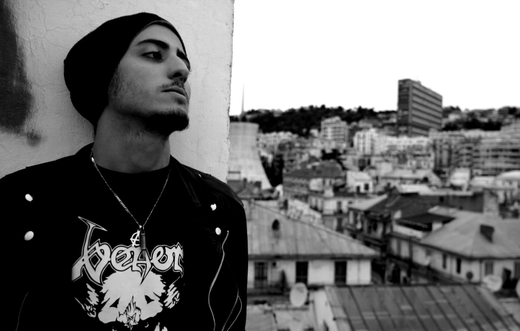 L'image montre un jeune homme sur un toit, regardant au loin. Il porte un t-shirt noir avec un motif graphique et une veste. Son look est accentué par un bonnet. En arrière-plan, on peut voir une vue de la ville avec des bâtiments et des toits, le tout dans des teintes en noir et blanc qui donne une atmosphère mélancolique. La scène évoque une ambiance urbaine et contemplative.