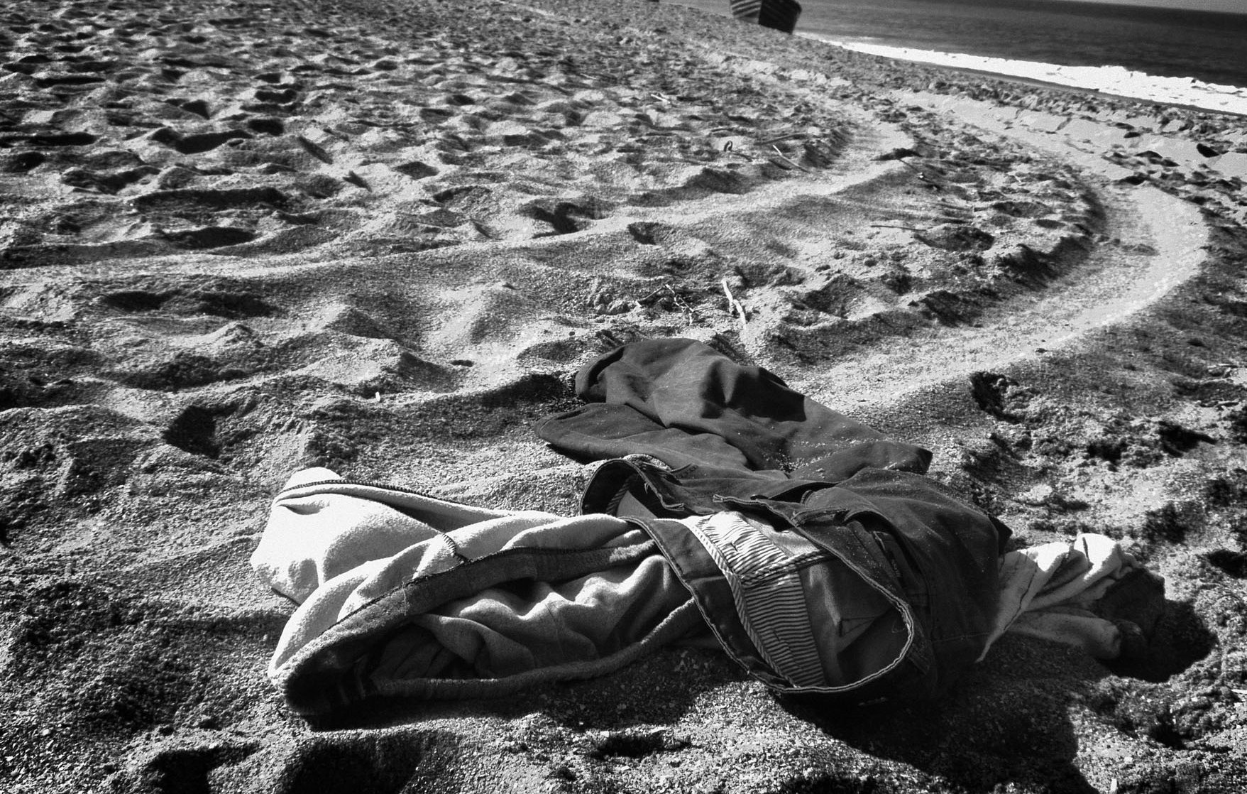 L'image montre un paysage de plage en noir et blanc, où des vêtements abandonnés sont éparpillés sur le sable. On peut voir des traces laissées par les pas dans le sable, créant un motif dans la texture. À l'arrière-plan, la mer se déroule tranquillement, et le ciel est clair. L'ensemble dégage une atmosphère de solitude et d'abandon.