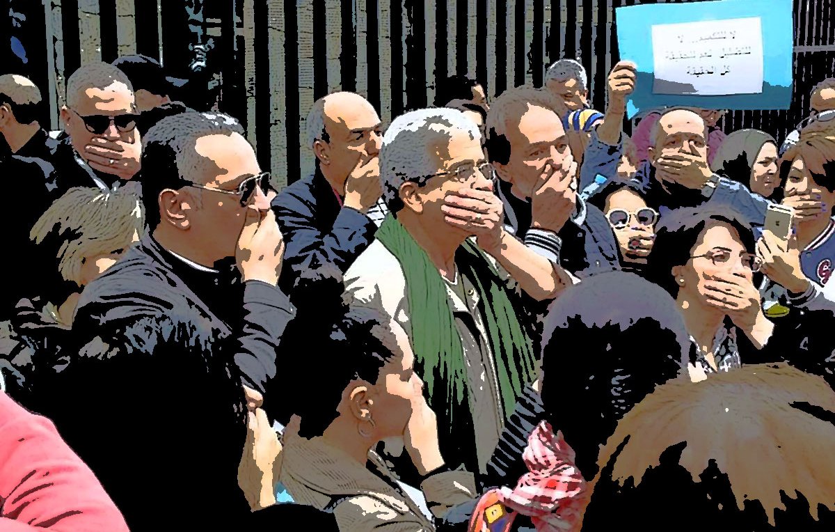 L'image montre un groupe de personnes réunies dans un cadre extérieur. Beaucoup d'entre elles se tiennent avec la main sur la bouche, semblant exprimer un silence ou protester. En arrière-plan, on aperçoit un panneau qui semble contenir un message. L'ambiance générale semble sérieuse et engagée, suggérant une forme de mobilisation ou de manifestation. Les participants semblent concentrés et solidaires.