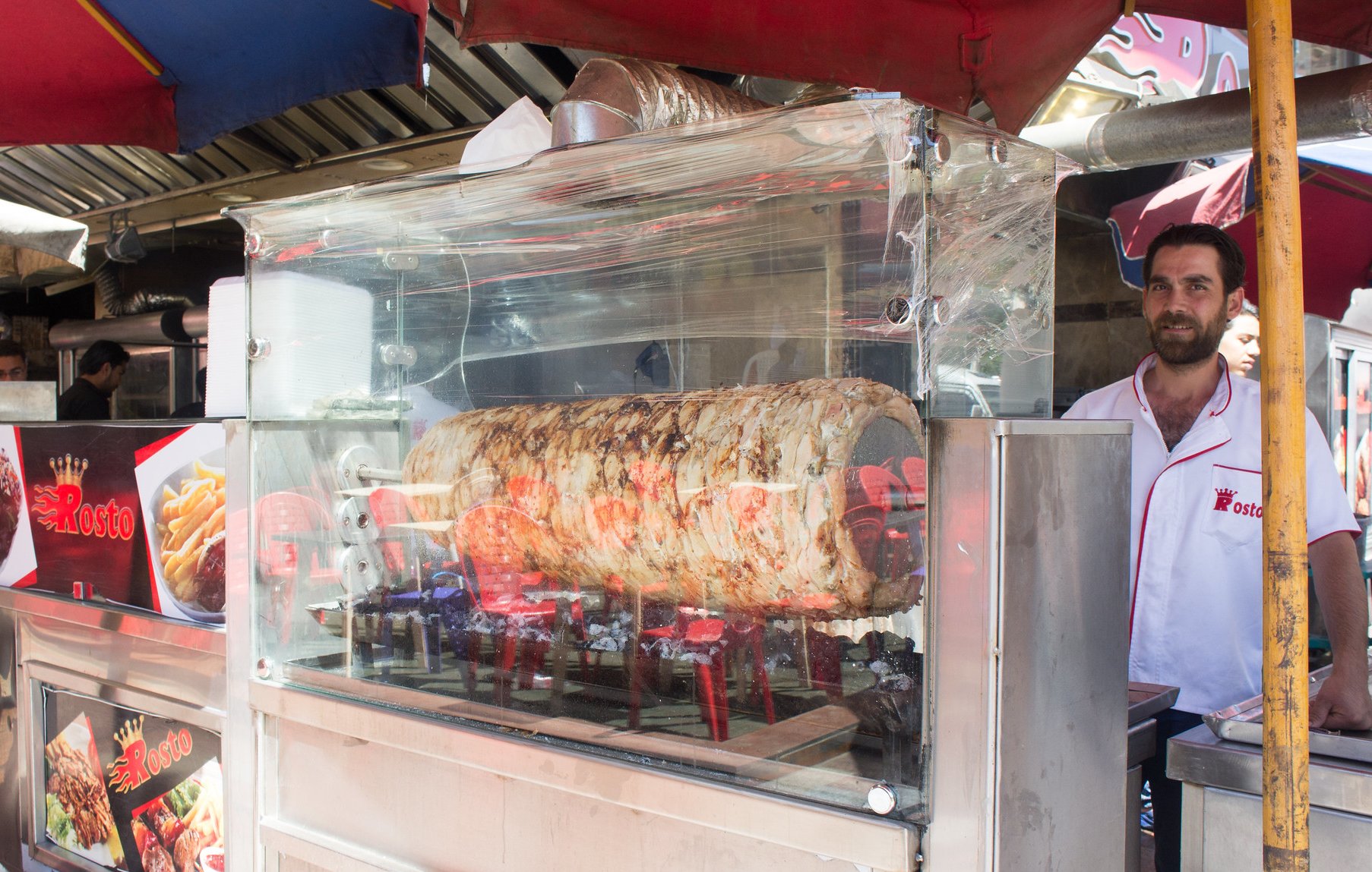 L'image montre un stand de nourriture de rue où un homme se tient derrière une vitrine en plastique transparente. À l'intérieur de la vitrine, il y a un grand morceau de viande en train de rôtir sur des braises, probablement un kebab ou un gyros. Le stand est couvert de parasols colorés, et il y a des affiches annonçant les plats proposés. L'ambiance semble vivante, typique des marchés animés de rues.