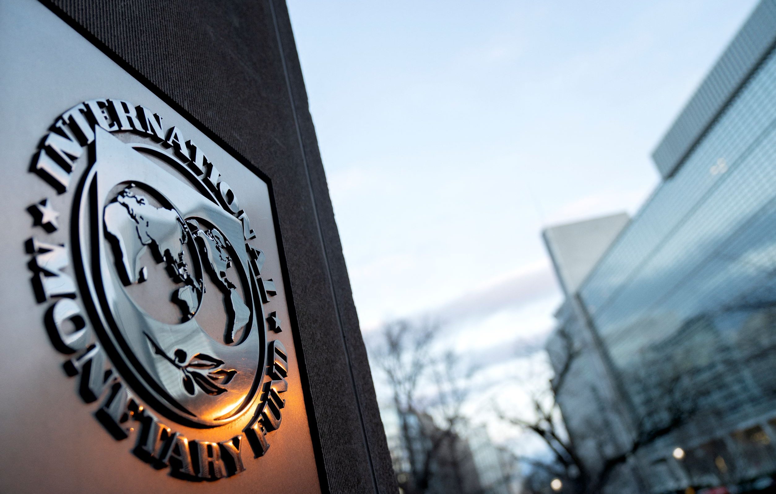 L'image montre le logo du Fonds monétaire international (FMI) en premier plan, avec un globe stylisé et des feuilles. En arrière-plan, on aperçoit un bâtiment moderne, probablement le siège du FMI, baigné dans une lumière douce du crépuscule. Le ciel est clair, avec des nuages légers. L'ensemble évoque une ambiance institutionnelle et internationale.