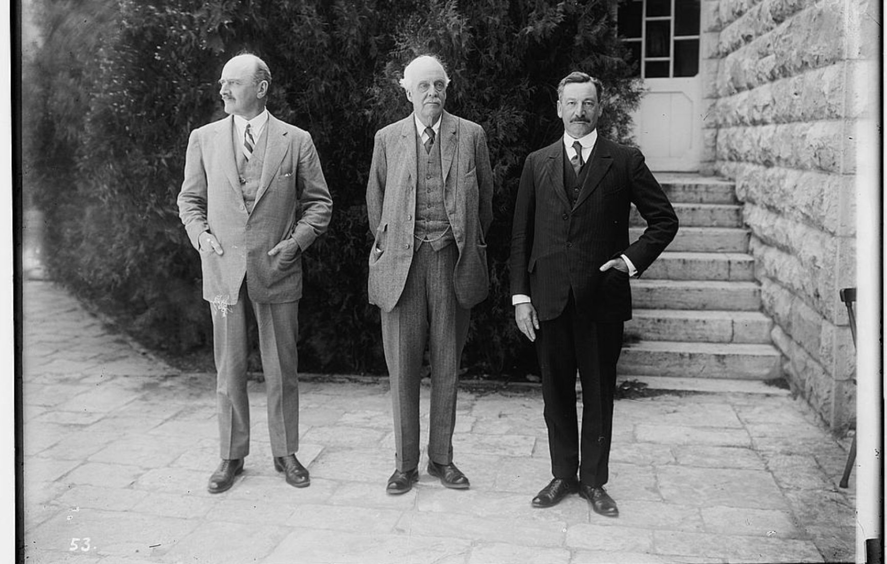 Trois hommes en costumes, debout devant un bâtiment avec des marches et des arbustes.
