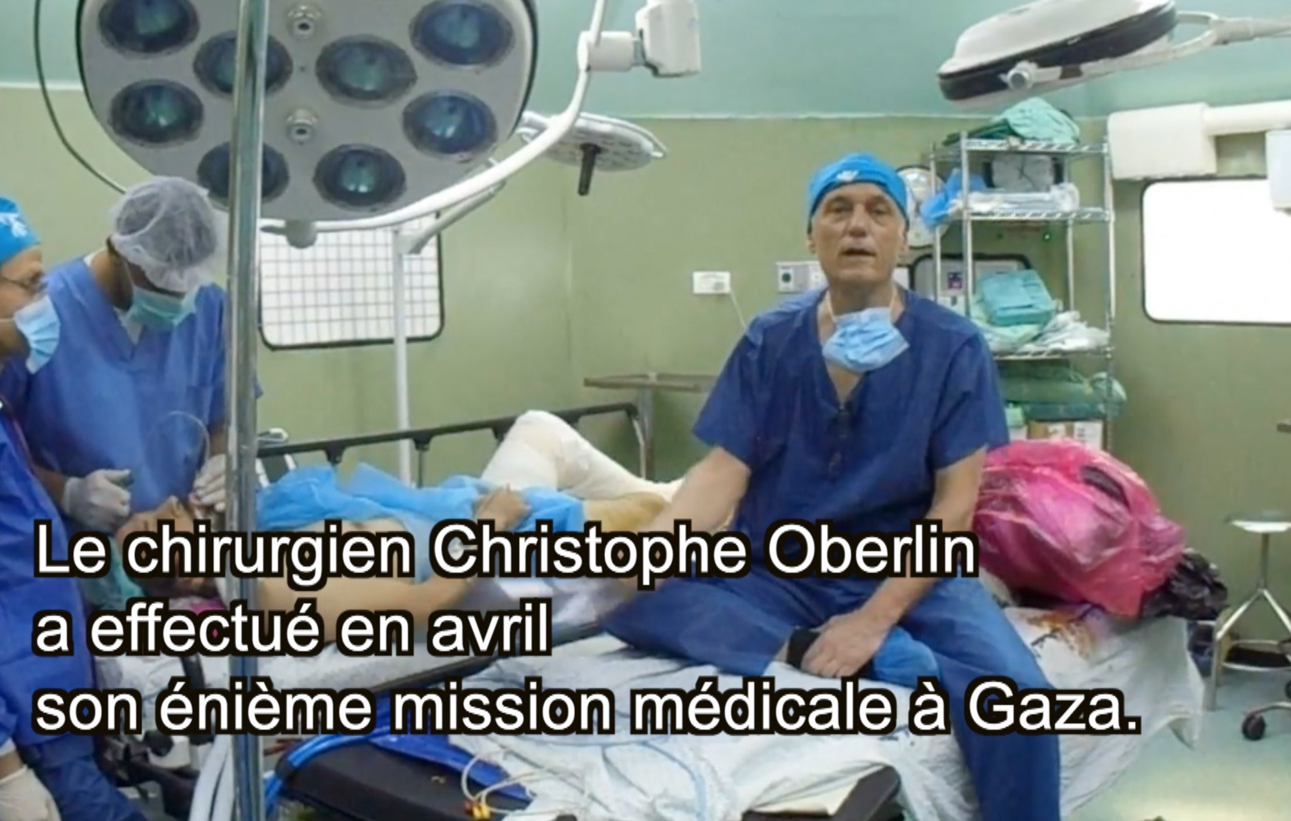 L'image montre une scène dans une salle d'opération. On y voit plusieurs professionnels de la santé en train de travailler. Ils portent des blouses et des masques de protection caractéristiques des environnements médicaux. Au centre, un patient est couché sur une table d'opération, tandis qu'un chirurgien, probablement Christophe Oberlin, s'adresse à la caméra. Des équipements médicaux, tels que des lampes et des instruments chirurgicaux, sont visibles dans la pièce. Le texte indique que cette intervention a eu lieu en avril lors d'une mission médicale à Gaza.