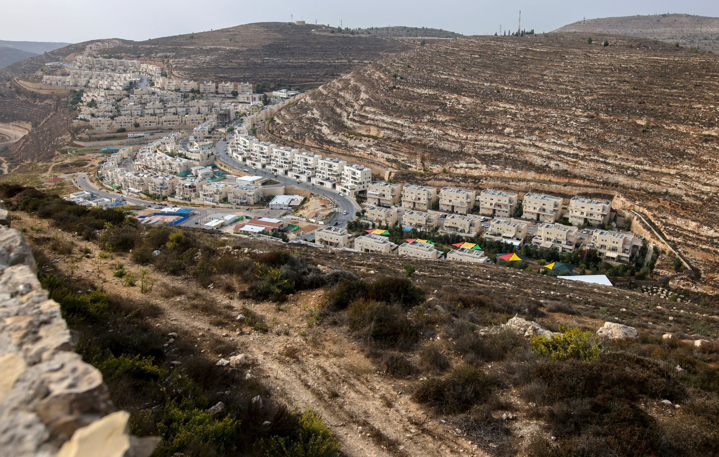 L'image montre un paysage vallonné avec des bâtiments résidentiels. On voit des maisons qui sont construites en terrasses sur les pentes de la colline, ainsi qu'une route qui serpente à travers l'urbanisation. La végétation est sparse et composée de buissons et d'herbes. Au fond, on aperçoit d'autres constructions, indiquant une zone développée et habitée. Le ciel est légèrement nuageux, ce qui donne une ambiance un peu grise à l'ensemble.