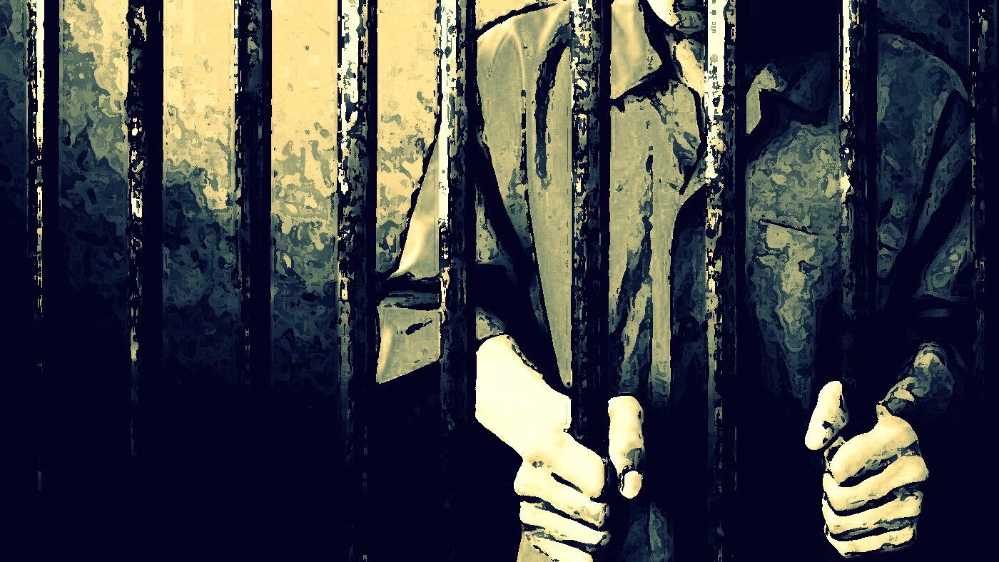 L'image montre des barreaux de prison, avec des mains qui s'agrippent à eux. Le fond est sombre et évoque une atmosphère de confinement. La personne derrière les barreaux est en partie visible, portant une chemise qui semble typique des uniformes de prison. L'éclairage crée des ombres, accentuant le sentiment d'isolement et de détention.
