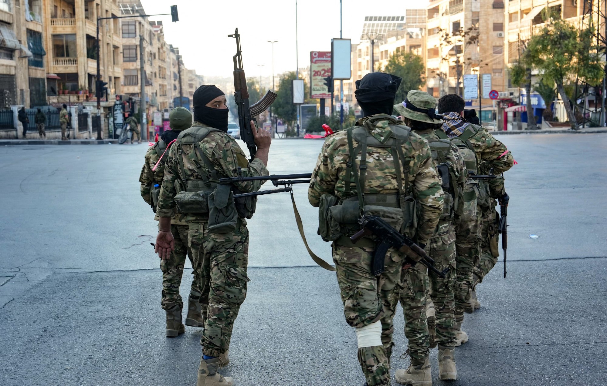 L'image montre un groupe de soldats armés marchant dans une rue urbaine. Ils portent des uniformes militaires et sont équipés d'armes. L'environnement semble urbain, avec des bâtiments en arrière-plan. L'ambiance est sérieuse, suggérant une situation potentiellement tendue. Les soldats portent également des masques, ce qui peut indiquer des préoccupations de sécurité.