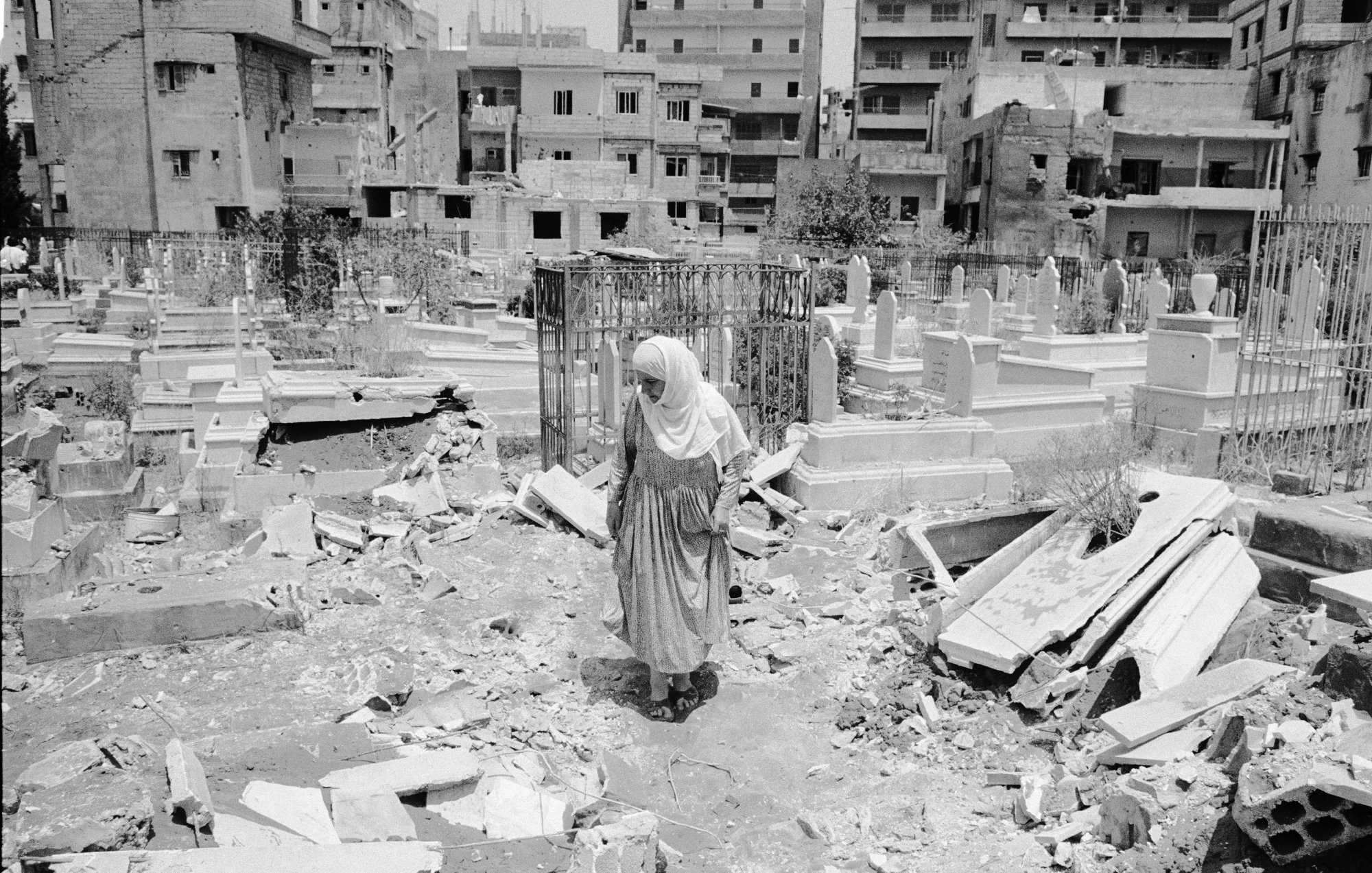 L'image montre une femme vêtue d'un long habit, marchant au milieu d'un cimetière. Le sol est désordonné, avec des débris éparpillés, ce qui suggère qu'il y a eu des destructions récentes dans la zone. En arrière-plan, on peut voir des bâtiments, probablement endommagés, qui entourent le cimetière. L'atmosphère paraît lourde, évoquant des sentiments de tristesse et de désolation.