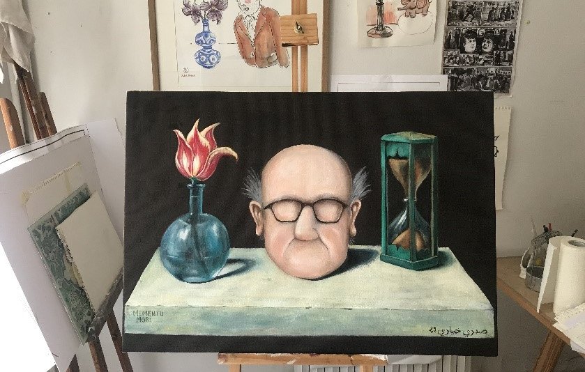 L'image montre une toile peinte, représentant un homme avec une tête large, des lunettes et des cheveux ébouriffés. Il est placé devant une table sur laquelle se trouvent un vase avec une fleur rouge, une sablier et un autre objet qui semble être une bouteille. En arrière-plan, on aperçoit des œuvres d'art accrochées au mur, ainsi qu'un chevalet. L'ensemble a une atmosphère artistique et quelque peu surréaliste.