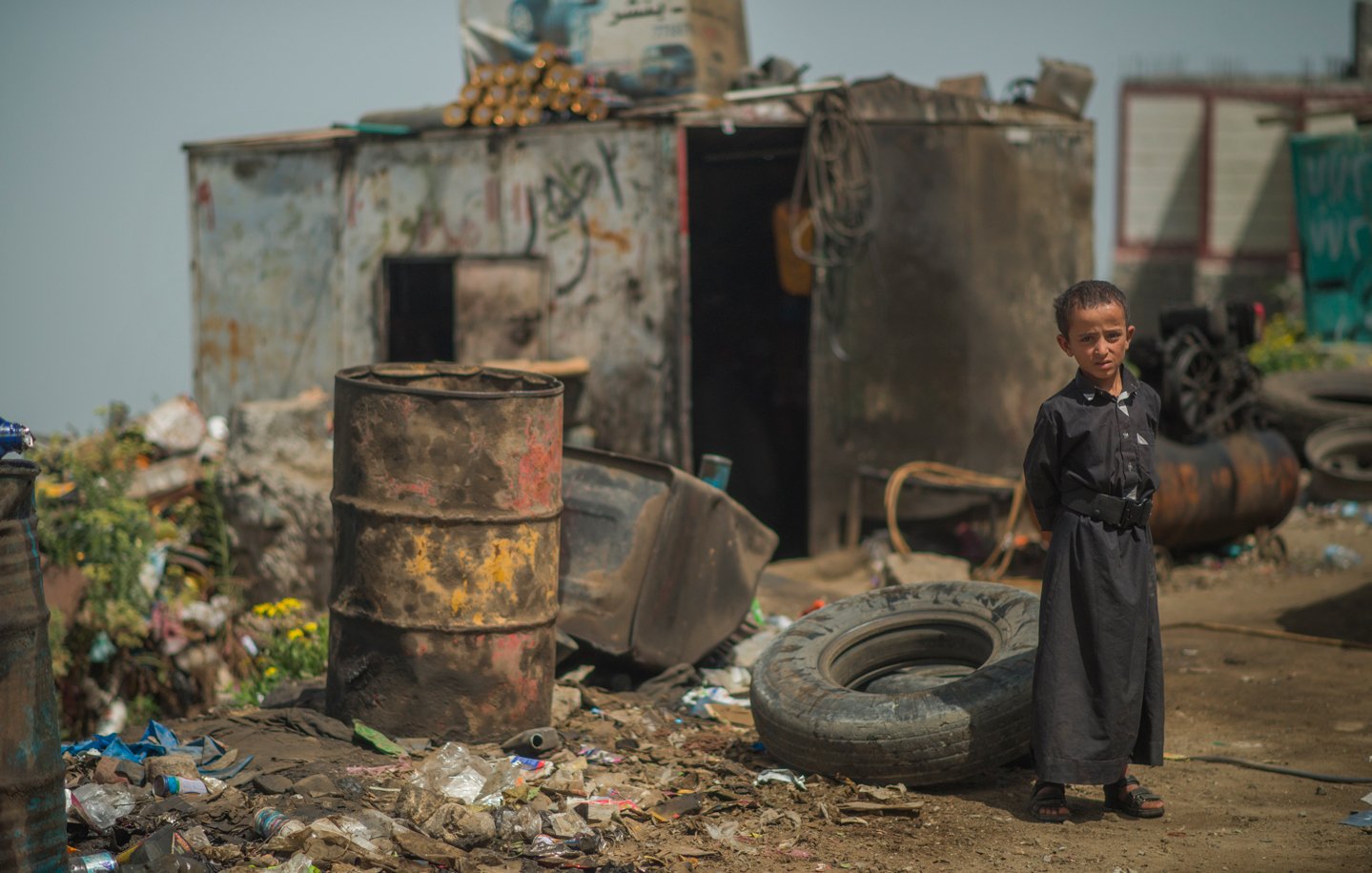 L'image montre un enfant debout dans un environnement désolé. Il porte des vêtements sombres, semblant se tenir près d'une zone remplie de débris, y compris des pneus usés et des barils. En arrière-plan, on aperçoit une structure rustique qui semble être une sorte d'abri. Le sol est sale, jonché de déchets, reflétant une atmosphère de pauvreté et de négligence. L'enfant a une expression sérieuse, ce qui souligne le caractère difficile de son environnement.