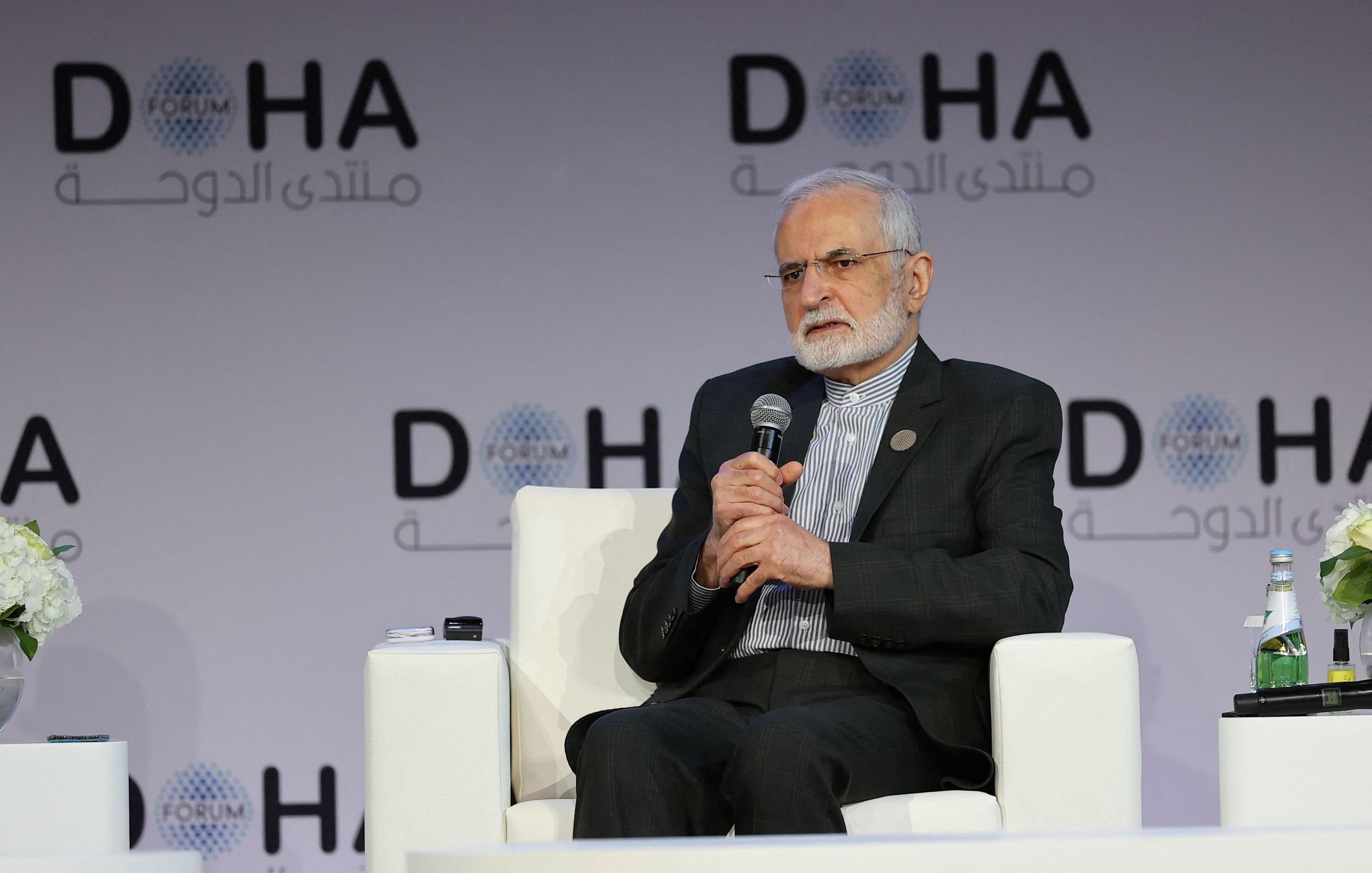 L'image montre un homme assis sur un fauteuil blanc, participant à une conférence ou un forum. Il porte une tenue formelle et semble être en train de s'exprimer ou d'écouter. En arrière-plan, on peut voir un logo avec le mot "DOHA" et d'autres inscriptions en arabe. Des fleurs et une bouteille d'eau sont également visibles sur la scène. L'atmosphère semble professionnelle et tranquille.