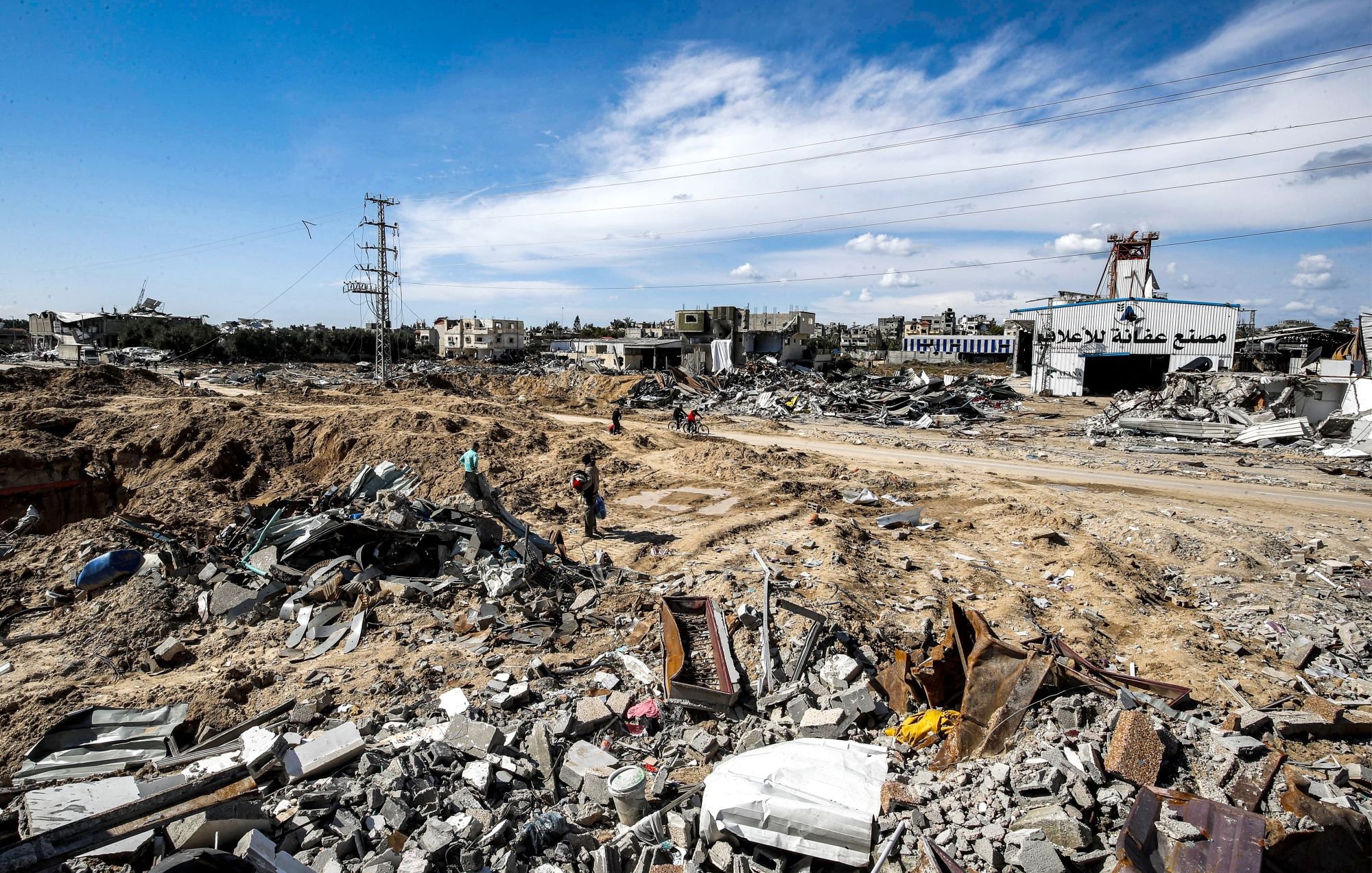 L'image montre un paysage dévasté, probablement dans une zone de conflit ou après une catastrophe. Le sol est jonché de débris, avec des morceaux de béton et de métal éparpillés. On peut apercevoir quelques personnes se déplaçant parmi les ruines, tandis qu'à l'arrière-plan, des bâtiments endommagés se dressent sous un ciel partiellement nuageux. Les lignes électriques sont visibles, indiquant une infrastructure perturbée. L'ambiance apparaît sombre et désolée, témoignant de la destruction qui a eu lieu.