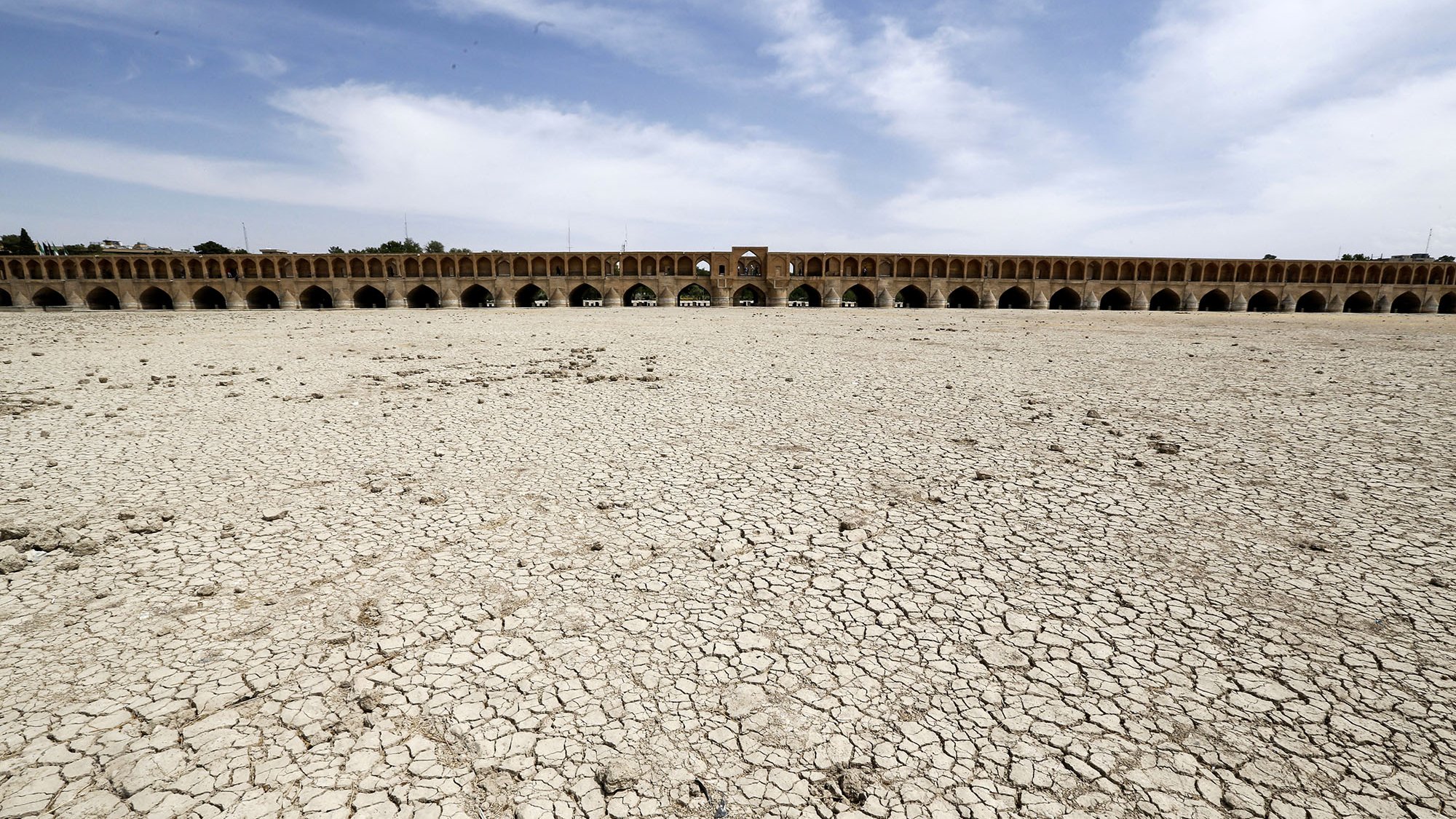 L'image montre un paysage aride avec des sols craquelés, indiquant une sécheresse sévère. À l'arrière-plan, on aperçoit un grand pont historique avec plusieurs arches. Le ciel est dégagé avec quelques nuages, et l'absence d'eau dans la zone suggère des conditions environnementales préoccupantes. Cette scène illustre les effets de la déshydratation sur un espace habituellement aquatique.