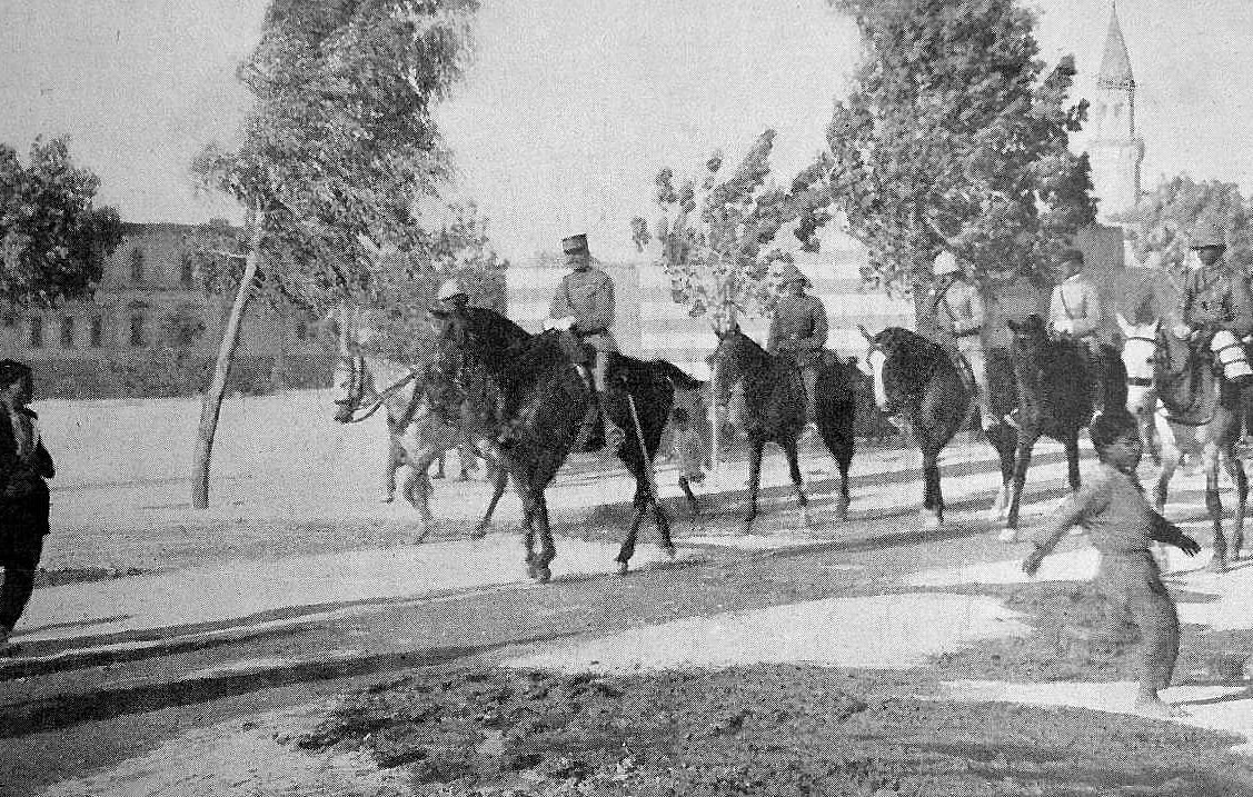 L'image montre un groupe de cavaliers, probablement des soldats, sur des chevaux, défilant sur une route. Il y a des arbres sur les côtés, et au premier plan, on peut voir une personne marchant sur le côté du chemin. À l'arrière-plan, des bâtiments et un clocher sont visibles, suggérant un environnement urbain. L'atmosphère semble historique, peut-être photographiée au début du XXe siècle.
