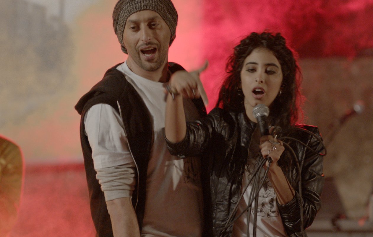 L'image montre des artistes en pleine performance musicale, avec une ambiance vibrante et colorée. Au centre, une femme chante dans un microphone, accompagnée d'un homme à ses côtés qui semble participer activement à la performance. En arrière-plan, d'autres musiciens se joignent à l'animation, entourés de lumière et de fumée, créant une atmosphère dynamique et festive. Les expressions des artistes indiquent de l'enthousiasme et de l'énergie.