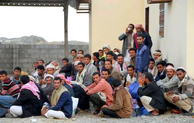 L'image montre un groupe de nombreux hommes assis au sol, semblant se regrouper près d'un bâtiment. Certains portent des vêtements traditionnels, tandis que d'autres ont des vêtements plus modernes. Ils semblent discuter ou écouter attentivement quelque chose. L'arrière-plan présente des murs en pierre et une construction simple. L'atmosphère semble calme, mais on peut sentir une certaine intensité dans leur rassemblement.