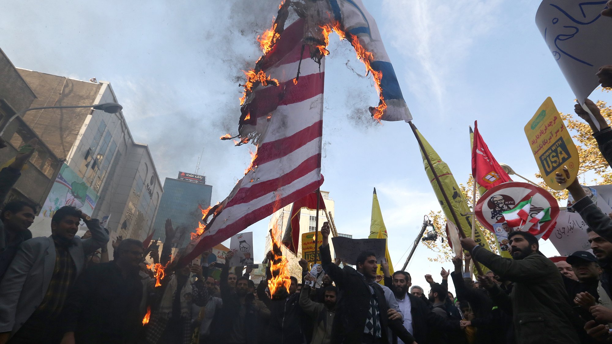 L'image montre une scène de manifestation où des personnes sont rassemblées pour protester. Au centre, un drapeau semble être en feu, ce qui attire l'attention des manifestants. Ils tiennent également des pancartes avec différents slogans et symboles. L'atmosphère est tendue, et on ressent un fort sentiment d'engagement parmi les participants. L'arrière-plan présente des bâtiments urbains, suggérant une grande ville.
