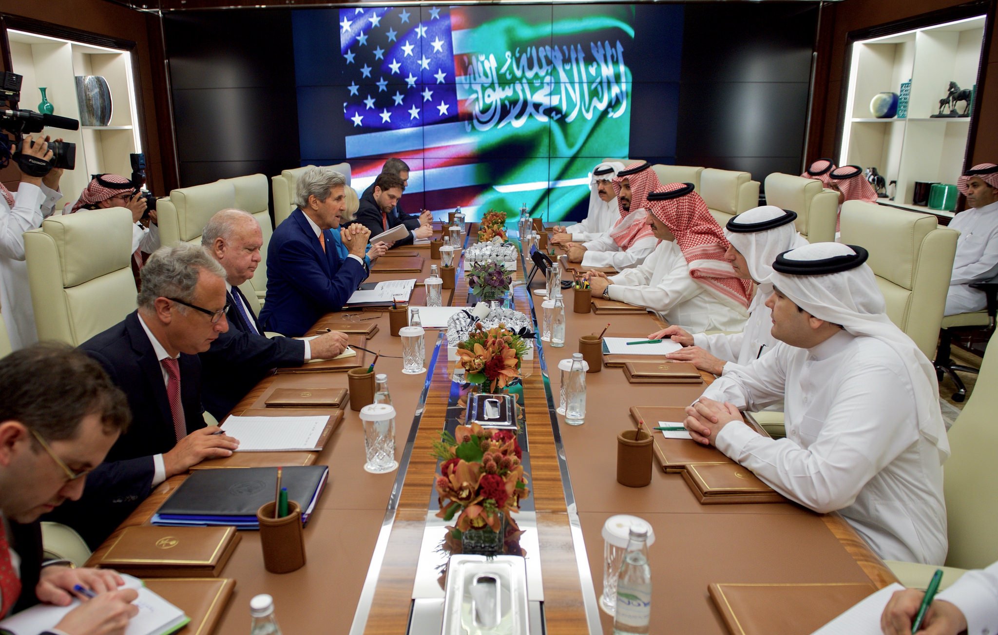L'image montre une réunion diplomatique dans une salle de conférence. On peut voir plusieurs personnes assises autour d'une longue table. À l'arrière-plan, il y a un écran affichant les drapeaux des États-Unis et d'Arabie saoudite. Les participants semblent être engagés dans une discussion sérieuse, avec des documents et des ordinateurs portables devant eux. L'atmosphère est formelle et professionnelle. Les membres de la réunion sont habillés en costumes et en vêtements traditionnels arabes.