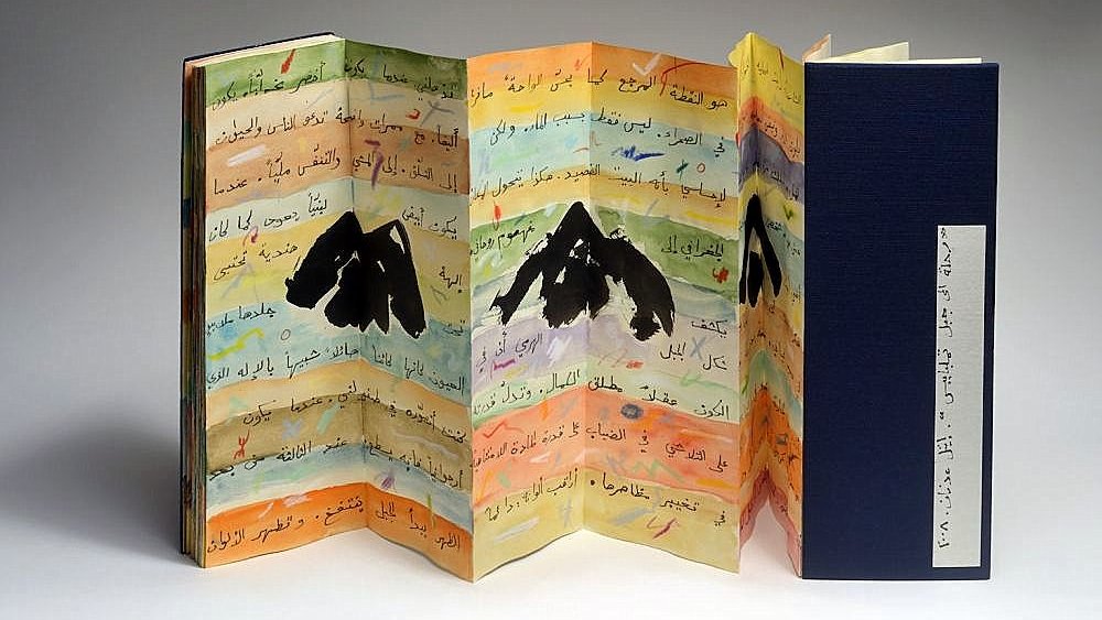 L'image montre un livre artistique aux pages colorées, présentant des illustrations de montagnes. Les pages sont pliées en accordéon et comportent des inscriptions en arabe. La palette de couleurs est variée, avec des nuances de jaune, bleu et orange, ajoutant une dimension vibrante à l'ensemble. Le livre est partiellement ouvert, révélant sa structure unique et sa conception créative. Une couverture noire est visible à droite, contrastant avec les couleurs vives des pages intérieures.