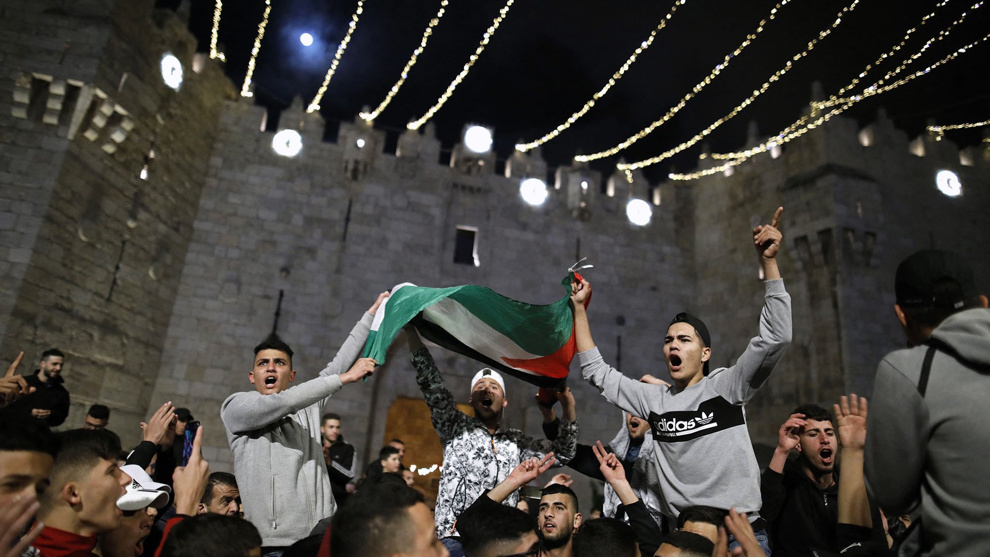 L'image montre un rassemblement nocturne devant un bâtiment ancien, probablement une fortification. Des personnes sont réunies en célébrant, certaines agitant un drapeau palestinien. L'atmosphère semble festive et dynamique, avec des lumières scintillantes qui illuminent la scène. Les expressions des participants retranscrivent une ambiance d'excitation et d'engagement.
