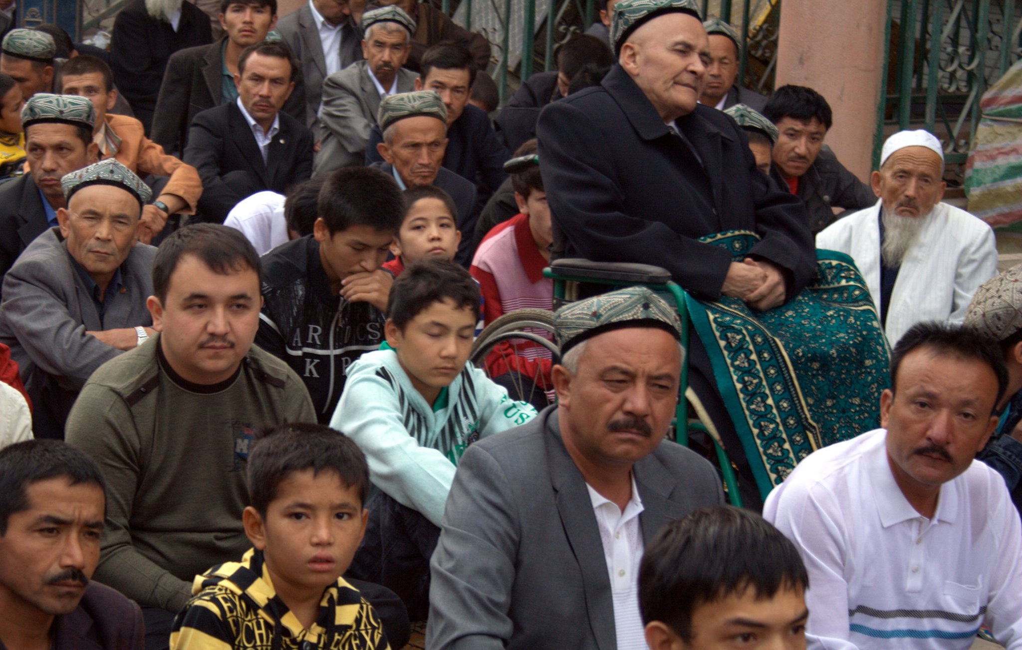 L'image montre un groupe de personnes assises, probablement lors d'un rassemblement ou d'une prière. On voit des hommes de différentes générations, certains portant des chapeaux traditionnels. Au centre, un homme âgé est assis sur une chaise, entouré de jeunes et d'adultes qui semblent attentifs. L'ambiance est sérieuse et respectueuse, reflétant un moment important pour la communauté.