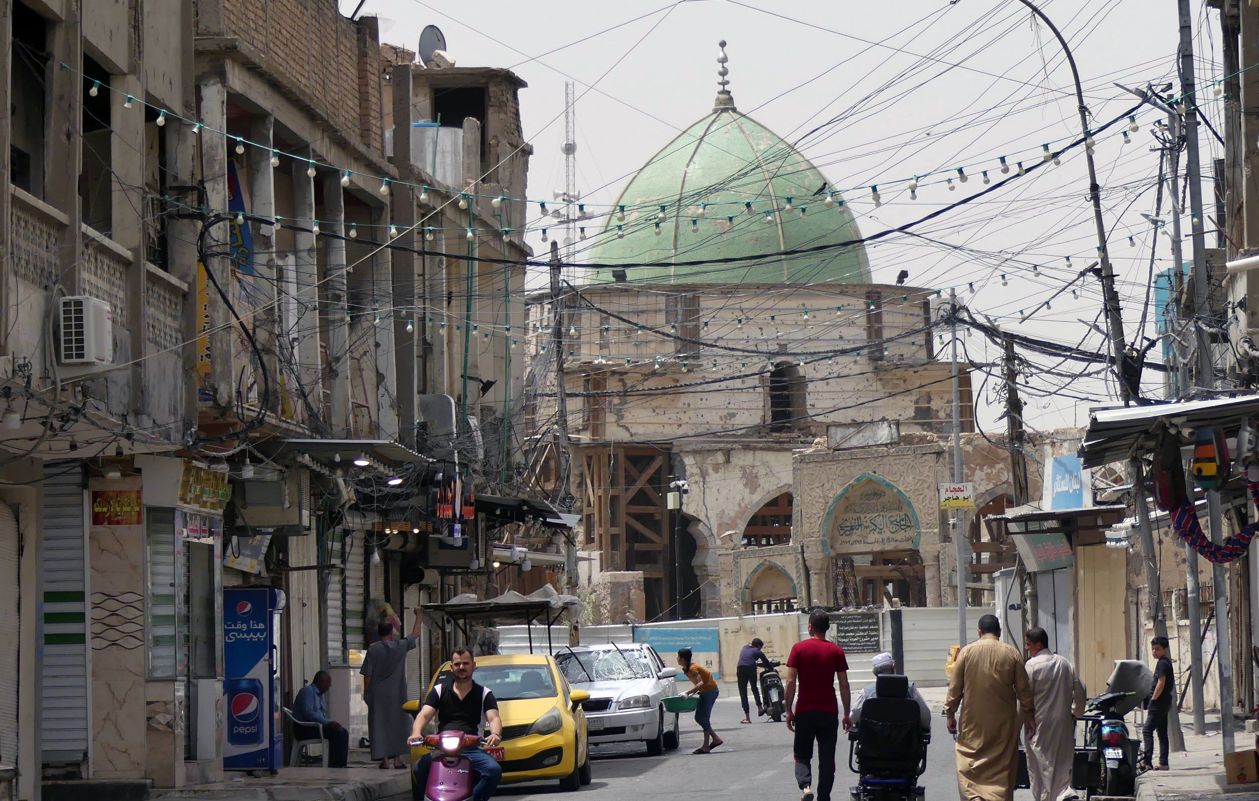 L'image montre une rue animée dans une ville. On y voit des bâtiments anciens avec des façades en pierre, certains étant en cours de rénovation. Au fond, une grande coupole verte domine la scène, indiquant la présence d'un bâtiment religieux. Les fils électriques sont visibles au-dessus de la rue. Des personnes marchent sur le trottoir, et une moto ainsi qu'une voiture jaune circulent. L'atmosphère semble à la fois traditionnelle et dynamique.
