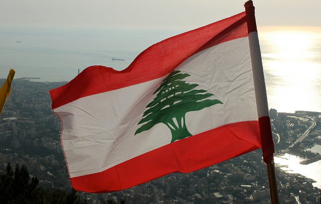 L'image montre le drapeau du Liban flottant au vent, avec un fond qui semble représenter une vue panoramique d'une ville côtière. Le drapeau se compose de trois bandes horizontales : rouge en haut et en bas, et une bande blanche au centre avec un grand cèdre vert, emblème national du Liban. L'horizon présente une mer scintillante, probablement au coucher du soleil, ce qui ajoute une ambiance paisible et naturelle à la scène.