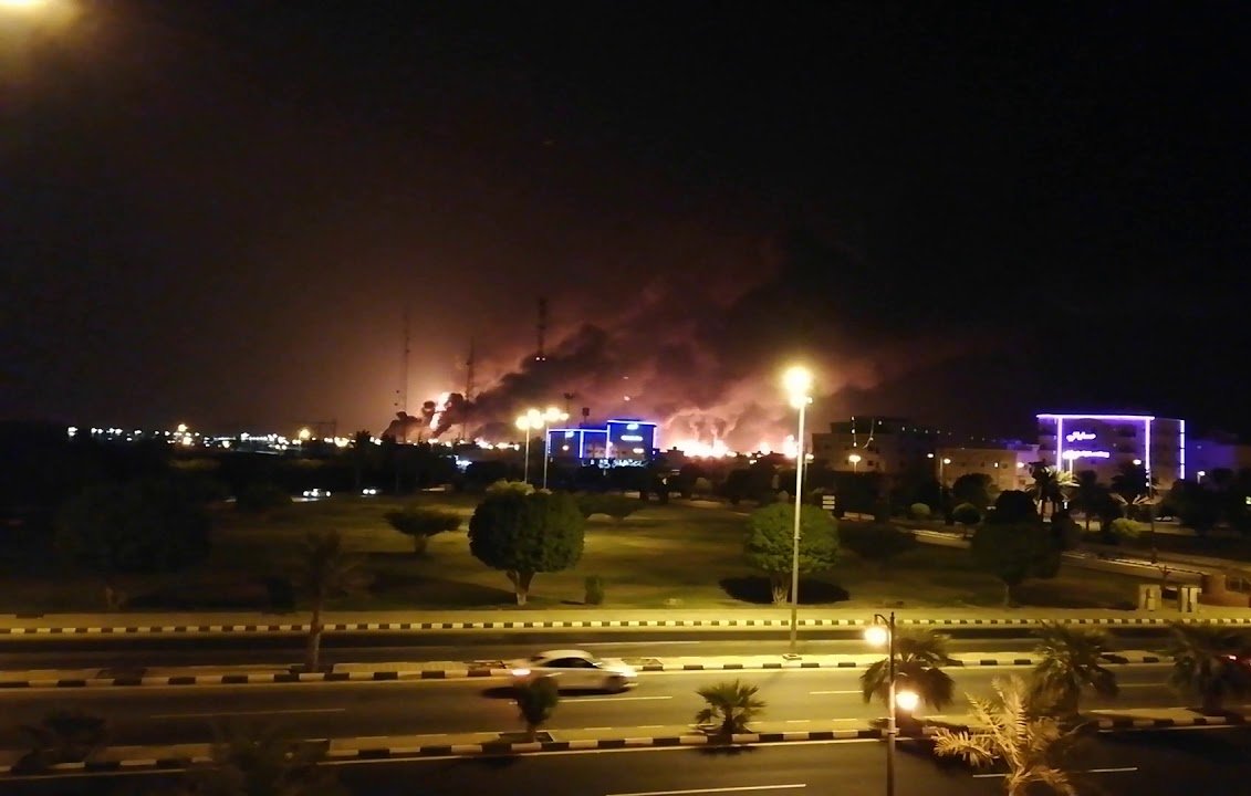 L'image montre une scène nocturne où l'on peut voir des nuages de fumée s'élevant dans le ciel, suggérant qu'il y a un incendie ou une explosion quelque part à l'horizon. Les lumières des bâtiments environnants sont visibles, créant un contraste avec l'obscurité de la nuit. La route devant est éclairée, avec quelques voitures qui passent. Des arbres et des espaces verts se trouvent également au premier plan, ajoutant une certaine profondeur à la scène.