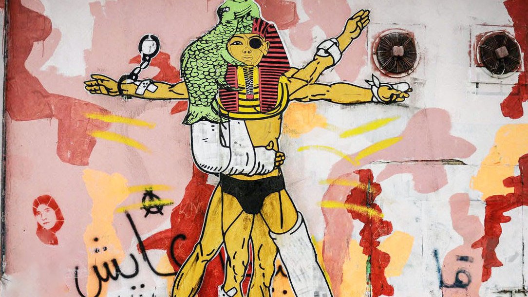 L'image représente une fresque murale colorée, incluant des éléments graphiques variés. Au centre, une figure humanoïde est stylisée avec des références à la culture égyptienne, incorporant des motifs inspirés des pharaons. Cette figure a plusieurs bras, symbolisant peut-être la force ou la multiplicités des rôles. Le fond est composé de formes abstraites et de couleurs vives, comme le rouge et le jaune, créant un contraste avec le personnage principal. On peut également apercevoir des inscriptions en arabe. L'ensemble véhicule une esthétique urbaine et contemporaine.