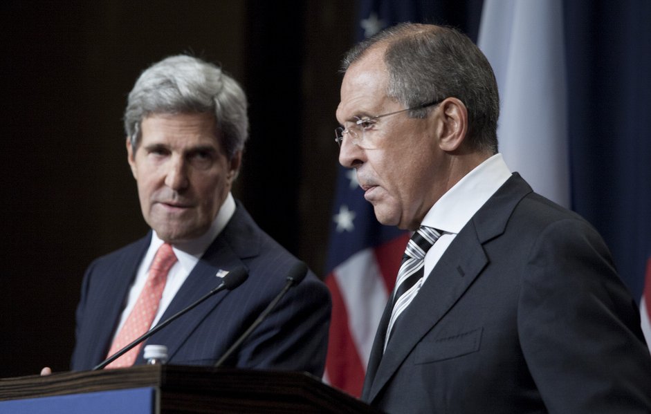 L'image montre deux hommes en tenue formelle, debout à un podium pendant une conférence de presse. L'un est à gauche, avec les cheveux gris et portant un costume bleu, et l'autre à droite, avec les cheveux foncés et portant un costume noir. Ils semblent être en train de discuter sérieusement, avec des drapeaux en arrière-plan, suggérant un contexte diplomatique ou de relations internationales.