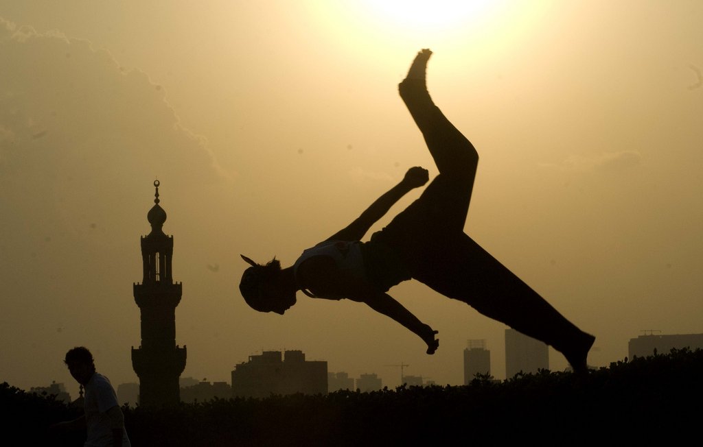 L'image montre une silhouette d'une personne effectuant un saut ou un mouvement acrobatique contre un fond de soleil couchant. À l'arrière-plan, on aperçoit une mosquée avec un minaret, ainsi que des bâtiments de la ville. L'éclairage crée un contraste fort entre la silhouette et le ciel lumineux, ajoutant une atmosphère dynamique et artistique à la scène.
