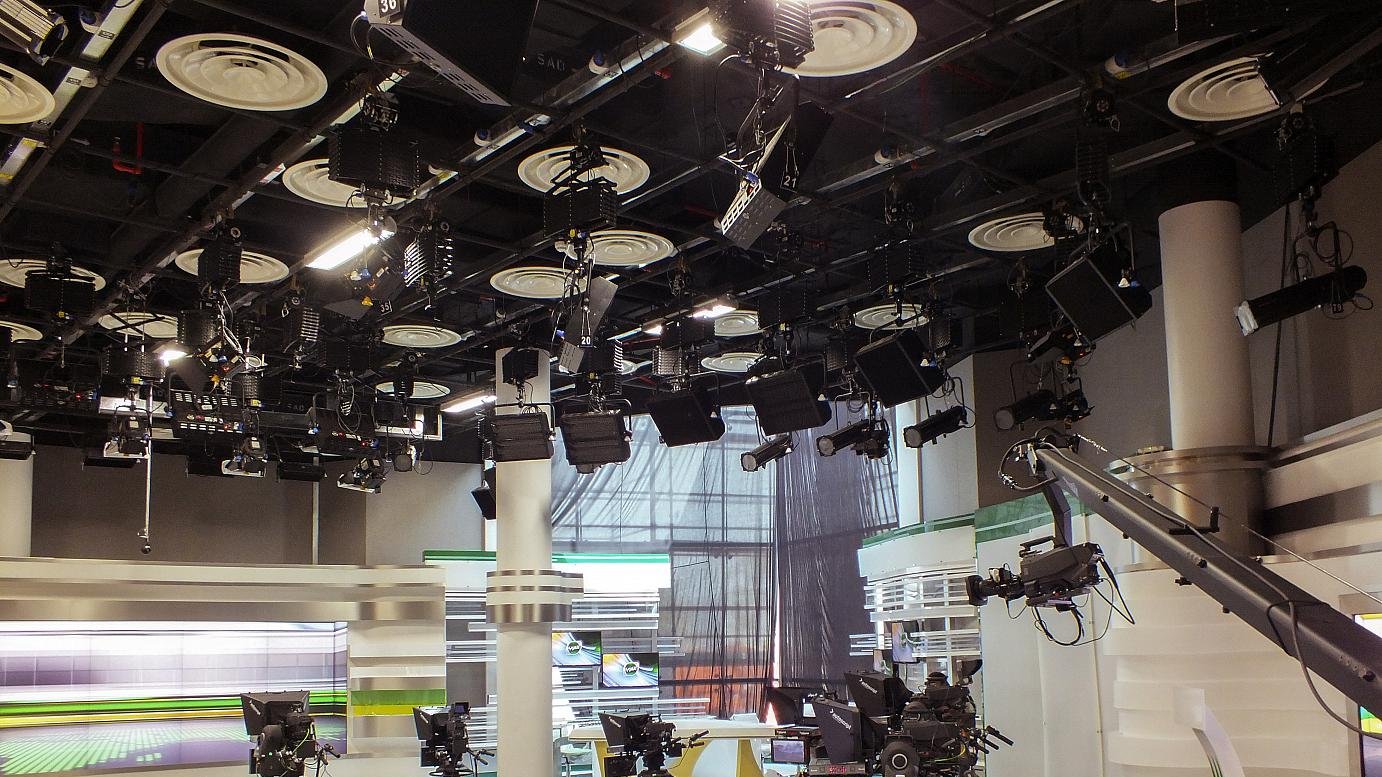 L'image montre un studio de télévision. On peut voir plusieurs caméras montées sur des tréteaux, prêtes à capturer des scènes. Le décor est moderne, avec des surfaces blanches et des accents de couleurs vives, notamment du vert. Des lumières suspendues au plafond éclairent le plateau, et il y a des écrans en arrière-plan, probablement utilisés pour afficher des informations ou des graphiques. L'ambiance semble professionnelle et organisée, typique d'un environnement de diffusion.