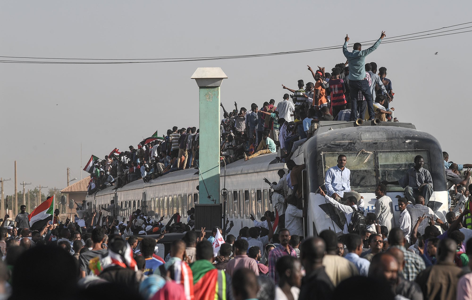 L'image montre une grande foule de personnes rassemblées autour et sur un train. Beaucoup de gens se tiennent sur le toit du train, tandis que d'autres s'accrochent aux côtés. L'atmosphère semble vibrante et énergique, probablement liée à une manifestation ou un événement important. En arrière-plan, on peut apercevoir des bâtiments et des infrastructures typiques d'une ville. La scène dégage un sentiment de solidarité et de détermination parmi les participants.