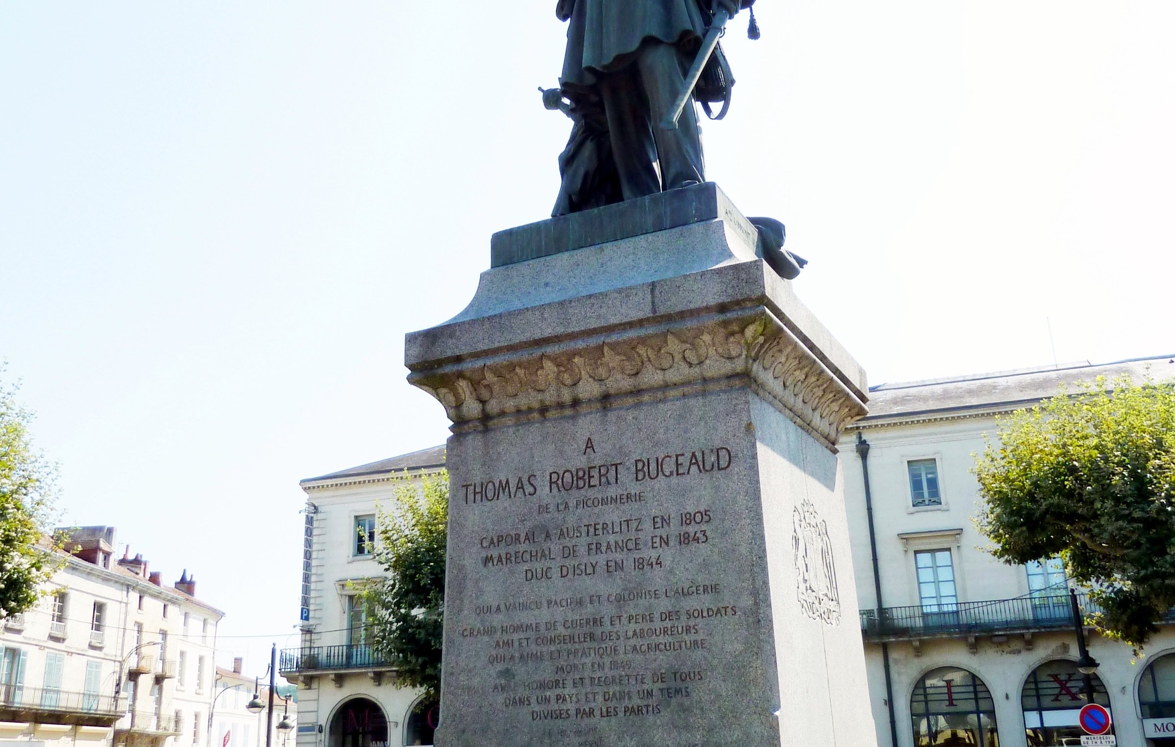 L'image représente une statue érigée à la mémoire de Thomas Robert Bugeaud. La statue est placée sur un socle en pierre portant une inscription détaillant ses accomplissements. Bugeaud est présenté en tenue militaire, tenant son épée. En arrière-plan, on peut voir une rue avec des arbres et des bâtiments, indiquant probablement un espace public. La lumière est claire, suggérant une journée ensoleillée.