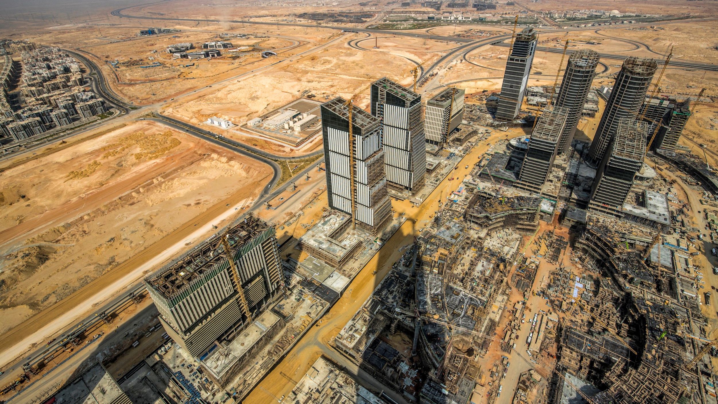 L'image montre une vue aérienne d'une région urbaine en développement, probablement dans un environnement désertique. On peut voir plusieurs gratte-ciels en construction, avec des structures modernes et des tours au milieu. Le paysage environnant est principalement désertique, avec peu de verdure, et des routes qui serpentent à travers la zone. La perspective donne une impression de grandeur et de modernité, tout en mettant en évidence les travaux d'infrastructure en cours.