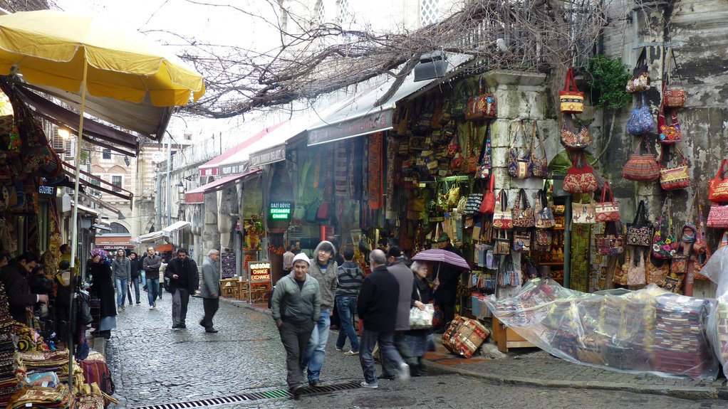 L'image montre une ruelle animée d'un marché. Les pavés sont humides, suggérant peut-être qu'il a récemment plu. Les étals sont remplis de sacs colorés et d'autres articles artisanaux, donnant une impression de vivacité et de diversité. Des gens se déplacent dans la rue, certains se protégeant sous des parapluies. Au-dessus, des branches nues d'arbres créent une atmosphère presque nostalgique, tandis que des auvents jaunes ajoutent une touche de couleur à la scène.