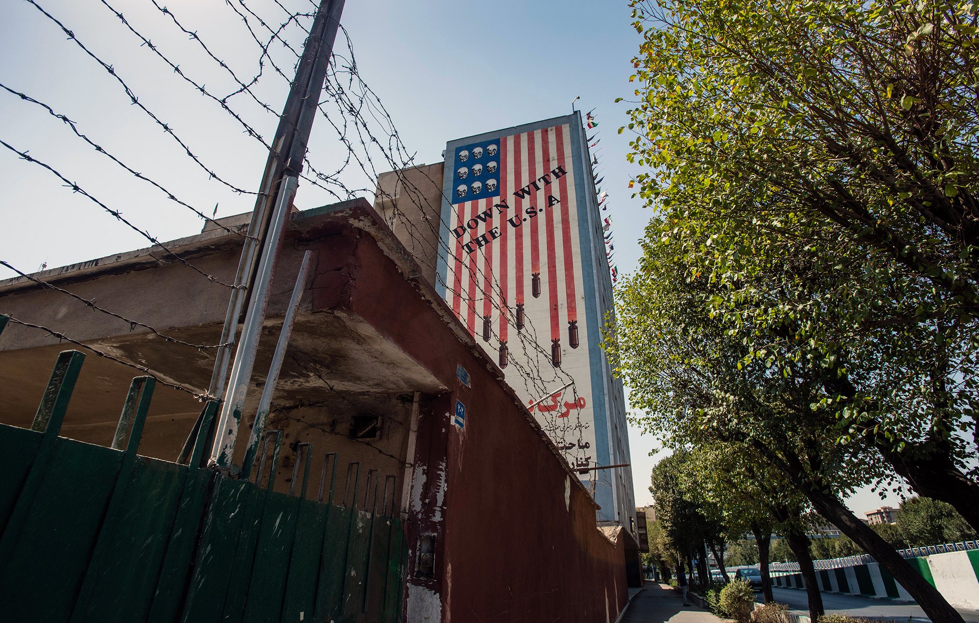 L'image montre un mur d'un bâtiment avec une grande peinture représentant un drapeau américain à rayures. Le mur est bordé par une clôture en barbelés, et on peut voir des arbres sur le côté. Le paysage est urbain, avec un chemin pavé qui suggère une rue à proximité. La composition évoque un contraste entre l'élément décoratif du drapeau et la présence de la clôture de sécurité.