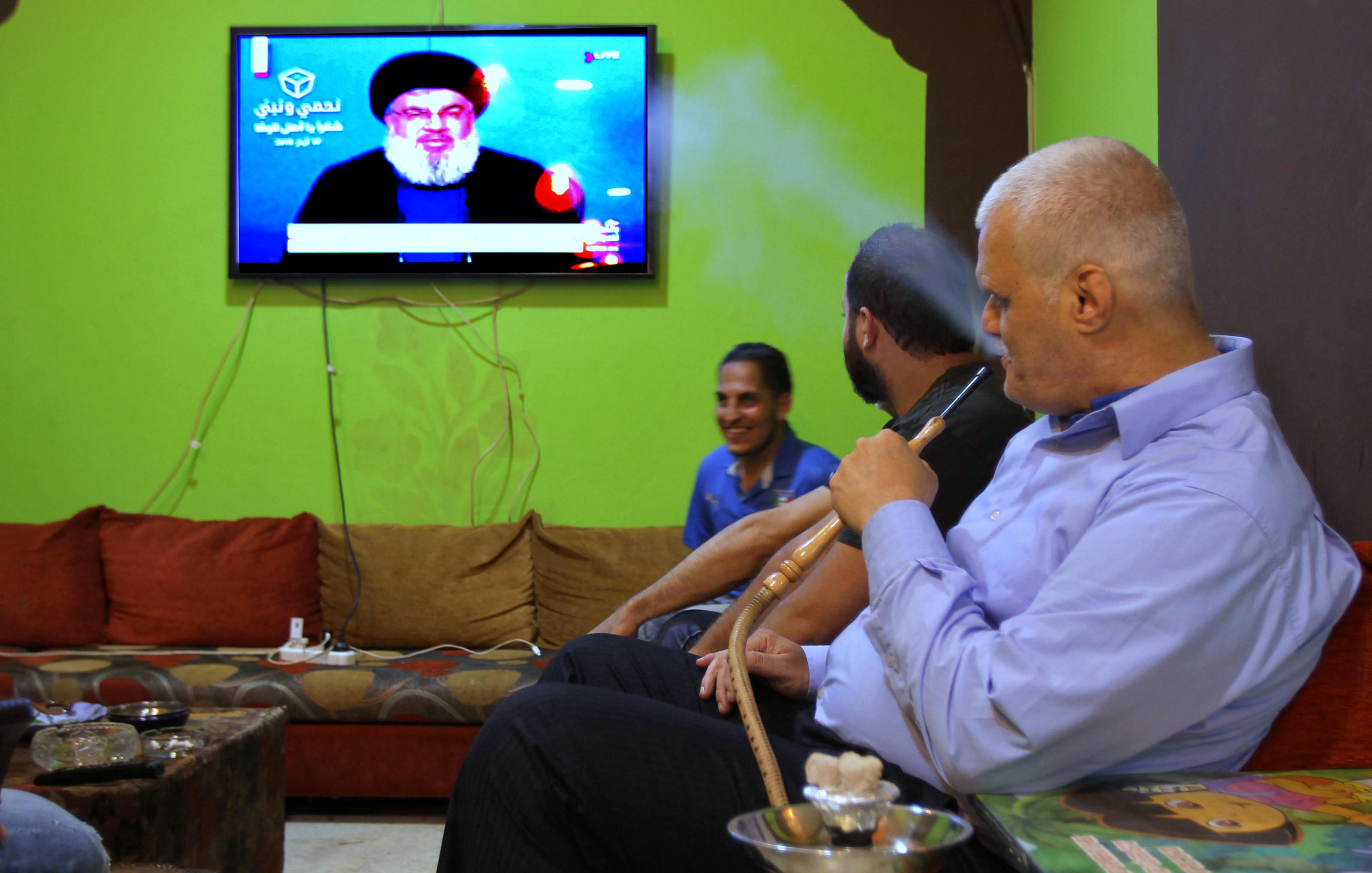 L'image montre un groupe d'hommes assis dans un salon avec des murs verts. Ils semblent regarder la télévision, où l'on peut voir un personnage à l'écran. L'un des hommes fume une pipe à eau, tandis que les autres sont engagés dans la conversation ou en observant l'écran. L'environnement est décontracté, avec des canapés confortables et des éléments de décoration simples.