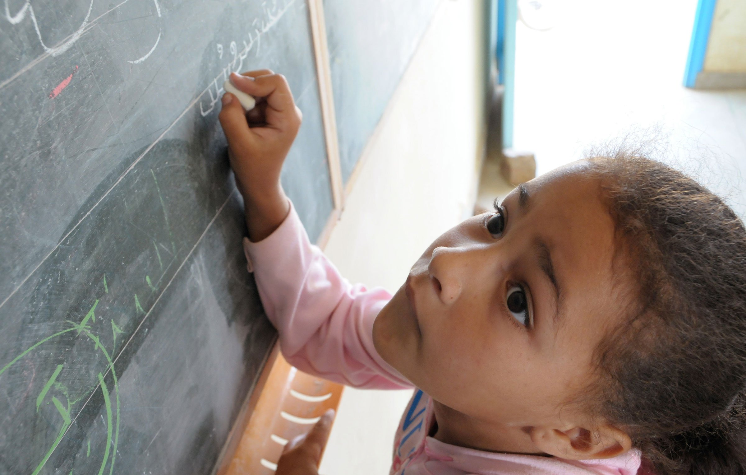 L'image montre une jeune fille penchée vers un tableau noir. Elle semble concentrée alors qu'elle écrit ou dessine quelque chose avec une craie. On peut voir des lettres ou des symboles tracés sur le tableau. La lumière entre par une fenêtre visible en arrière-plan, créant une ambiance chaleureuse. La fille porte un sweat à capuche rose et a les cheveux bouclés.