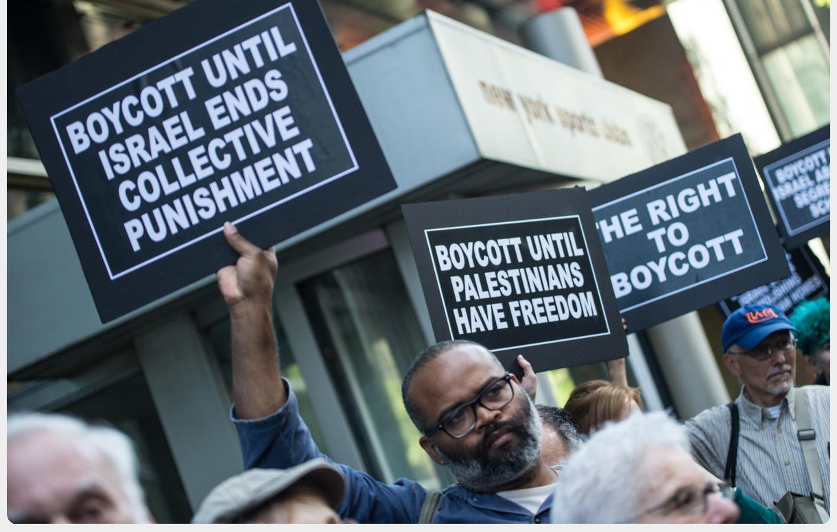 L'image montre une manifestation avec des personnes tenant des pancartes. Les pancartes portent des messages appelant au boycott jusqu'à ce qu'Israël mette fin à des mesures de punition collective et jusqu'à ce que les Palestiniens aient la liberté. On peut voir des participants de différents âges, tous unis par des revendications similaires. L'arrière-plan indique un environnement urbain avec des bâtiments.