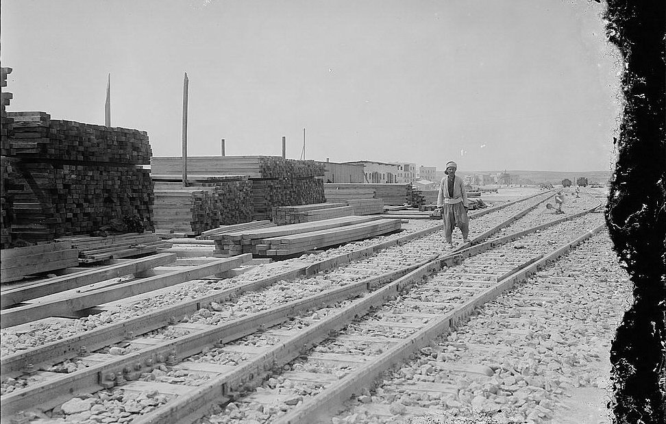 L'image montre une scène industrielle avec des voies de chemin de fer se dirigeant vers l'horizon. Sur le côté gauche, il y a des tas de planches de bois empilées, ce qui suggère une activité de construction ou de stockage. Un homme, probablement un travailleur, marche le long des rails. En arrière-plan, on aperçoit des bâtiments, donnant l'impression d'un site en pleine activité. Le ciel est dégagé, et la lumière est vive, typique d'une journée ensoleillée. L'atmosphère générale évoque une époque passée, avec une ambiance de travail acharné.