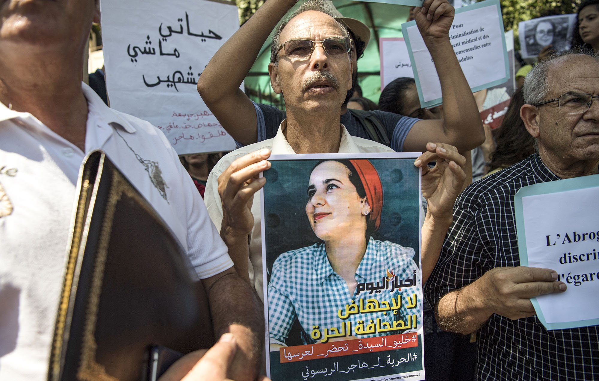 L'image montre un groupe de personnes rassemblées pour une manifestation. Au premier plan, un homme tient une affiche qui représente une femme avec un sourire, probablement une figure symbolique pour le mouvement. Les participants, majoritairement des adultes, tiennent également des pancartes avec des slogans en arabe et en français, indiquant leur lutte ou leur soutien à une cause, sans doute liée à des droits ou à une injustice. L'atmosphère semble déterminée et engagée.