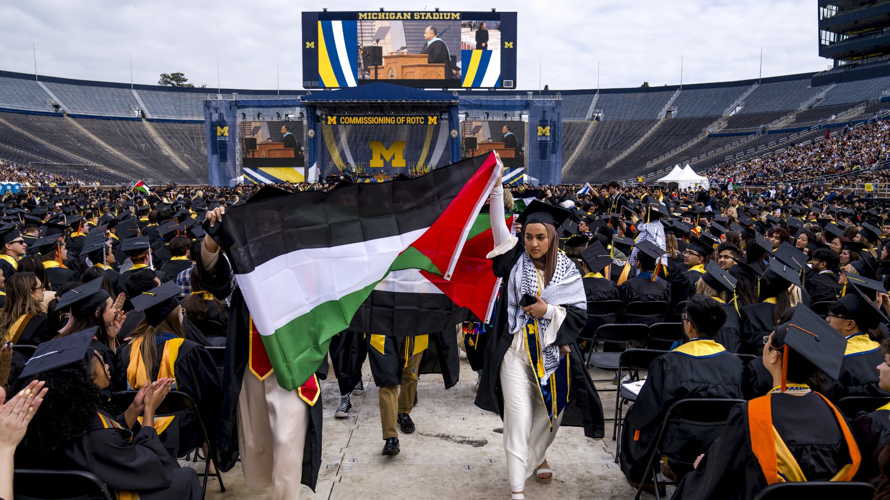 L'image montre une cérémonie de remise de diplômes qui se déroule dans un grand stade, probablement le Michigan Stadium. Des diplômés portant des toges et des chapeaux de cérémonie sont assis en rangées. Au premier plan, deux personnes tiennent des drapeaux : l'un est le drapeau palestinien et l'autre semble être un drapeau avec des couleurs distinctives. À l'arrière-plan, un grand écran affiche un orateur. L'ambiance est festive et solennelle, typique des cérémonies de graduation.