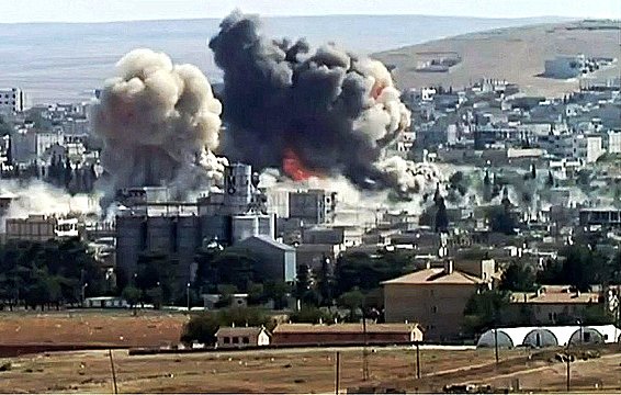 L'image montre un paysage urbain après une explosion, avec d'importants nuages de fumée et des décombres. Des bâtiments sont partiellement détruits, et l'atmosphère est sombre en raison de la fumée épaisse. Le sol semble être rural, avec des champs et quelques structures en arrière-plan. Cette scène évoque des conséquences dramatiques d'un conflit ou d'une attaque.