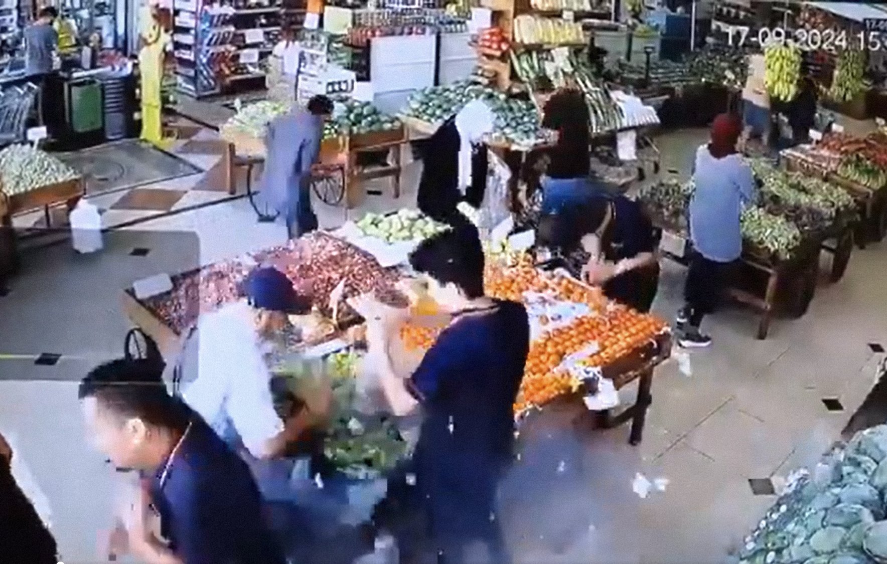 L'image montre une scène animée à l'intérieur d'un marché ou d'une épicerie. On peut voir plusieurs personnes qui interagissent autour de tables chargées de fruits et légumes. L'ambiance semble occupée, avec diverses personnes, certaines portant des sacs, en train de choisir des produits. Les étals sont colorés et bien garnis, suggérant une variété de produits frais disponibles. L'environnement paraît lumineux et accueillant.