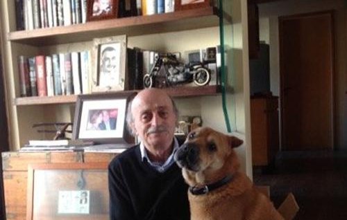 L'image montre un homme assis sur le sol, avec un chien à ses côtés. L'homme porte un pull noir et a des cheveux grisonnants. Le chien, de taille moyenne, a un pelage brun et semble calme. En arrière-plan, on peut voir une bibliothèque remplie de livres, ainsi que quelques objets personnels comme des cadres photo et des souvenirs. L'ambiance de la pièce paraît chaleureuse et accueillante.