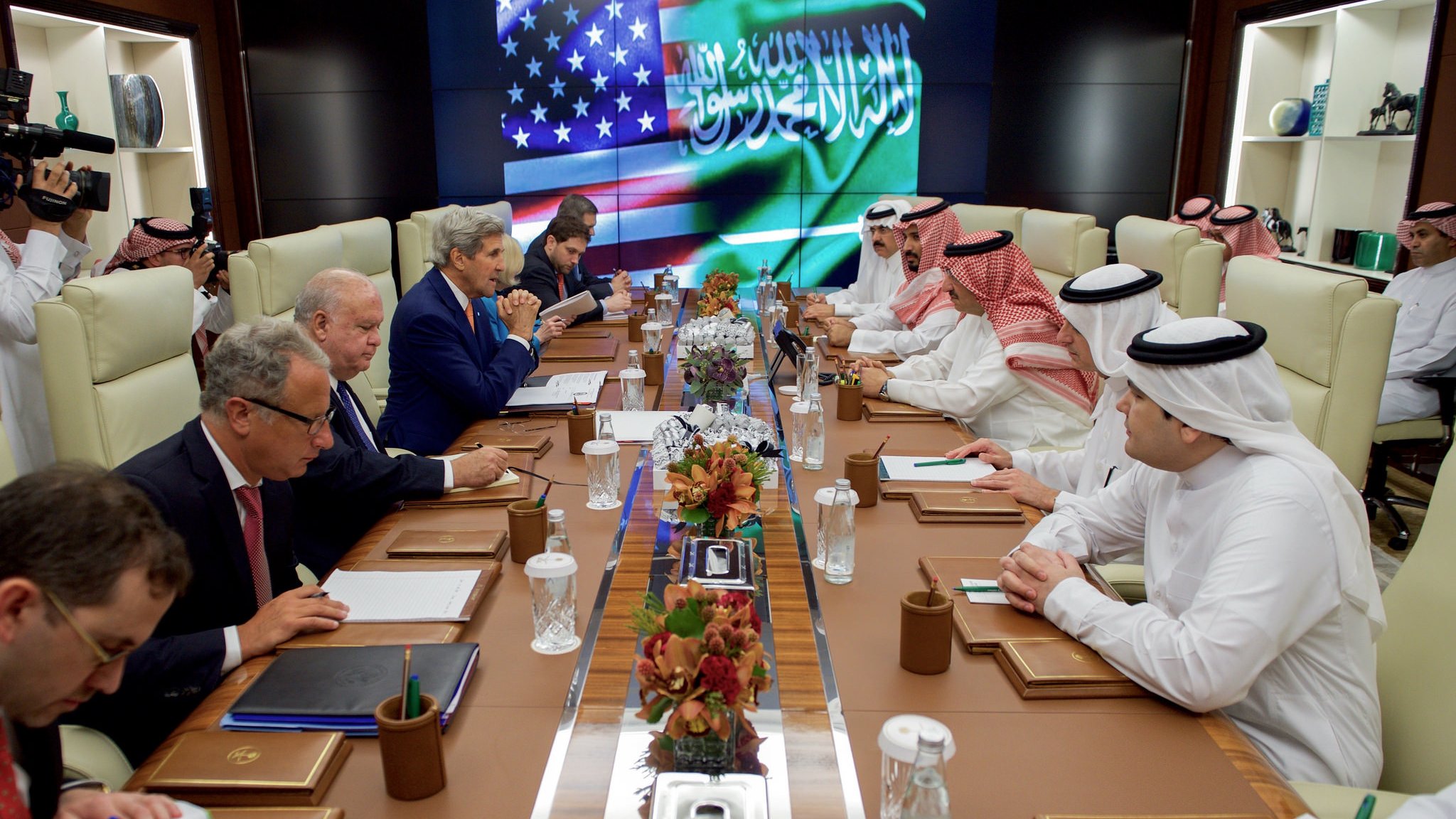 L'image montre une réunion diplomatique dans une salle de conférence. On peut voir plusieurs personnes assises autour d'une longue table. À l'arrière-plan, il y a un écran affichant les drapeaux des États-Unis et d'Arabie saoudite. Les participants semblent être engagés dans une discussion sérieuse, avec des documents et des ordinateurs portables devant eux. L'atmosphère est formelle et professionnelle. Les membres de la réunion sont habillés en costumes et en vêtements traditionnels arabes.
