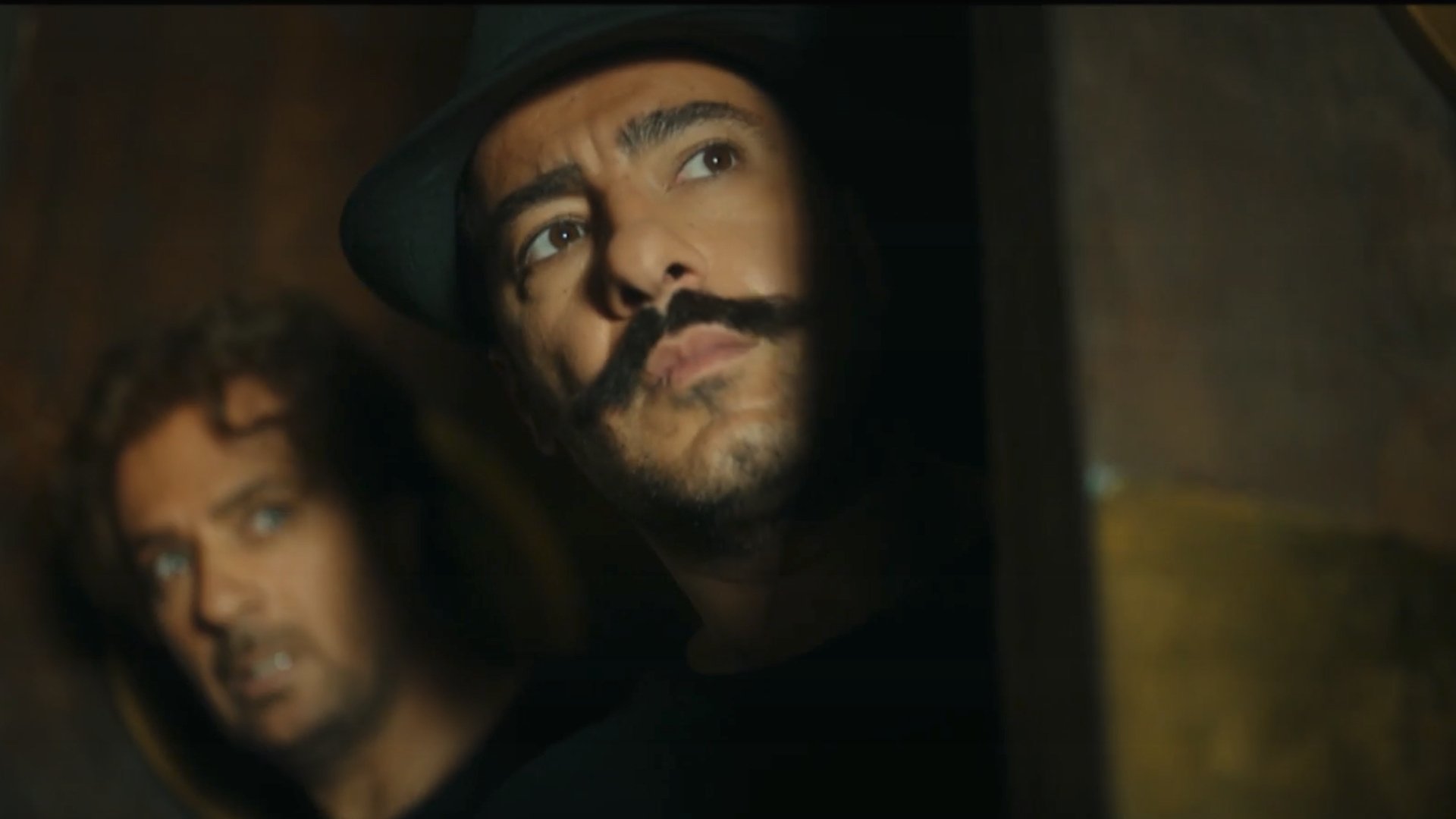 L'image montre deux hommes. L'un d'eux a une moustache prononcée et porte un chapeau, tandis que l'autre a les cheveux bouclés et semble inquiet. Ils regardent tous les deux vers la droite, comme s'ils étaient en train d'espionner ou de guetter quelque chose. L'éclairage est sombre, ce qui ajoute une atmosphère tendue et mystérieuse à la scène.