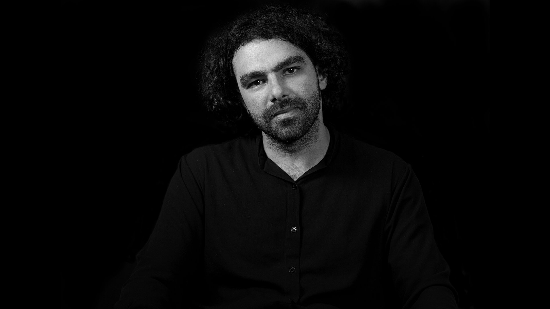 L'image montre un homme assis, regardant directement vers l'objectif. Il a des cheveux bouclés et une barbe, et il porte une chemise noire. Le fond est noir, ce qui crée un contraste net avec son visage éclairé. Son expression est neutre, suggérant une certaine introspection ou réflexion.