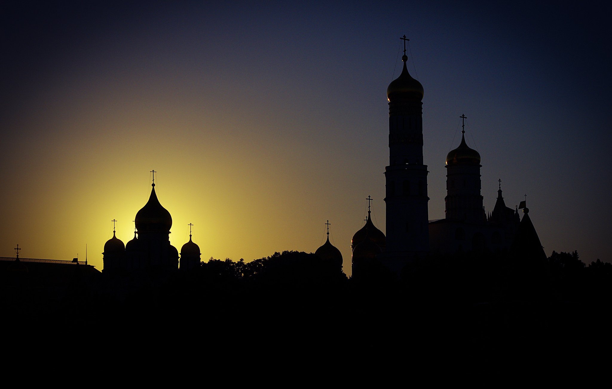 L'image présente un paysage urbain au crépuscule, où plusieurs structures architecturales se dressent en silhouette contre un ciel dégradé de couleurs. On peut apercevoir des dômes de style traditionnel, typiques de certaines églises, ainsi que des tours avec des croix au sommet. L'éclairage doux du soleil couchant crée un contraste accentué entre les ombres des bâtiments et le ciel lumineux, apportant une atmosphère calme et contemplative à la scène.